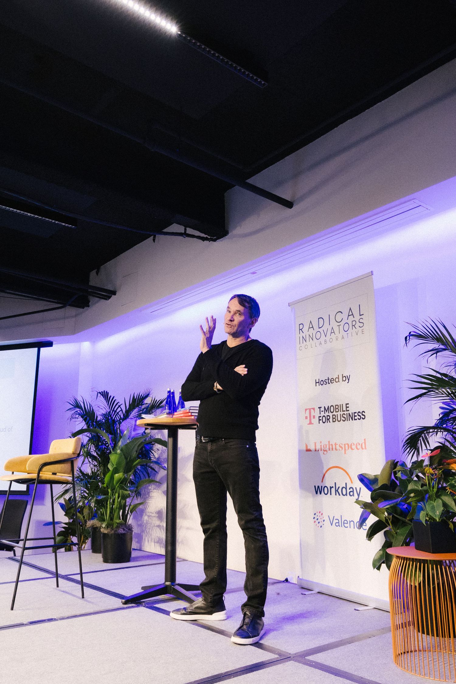 Speaker presents on stage with purple backlit wall and tropical plants at Radical Partners event.