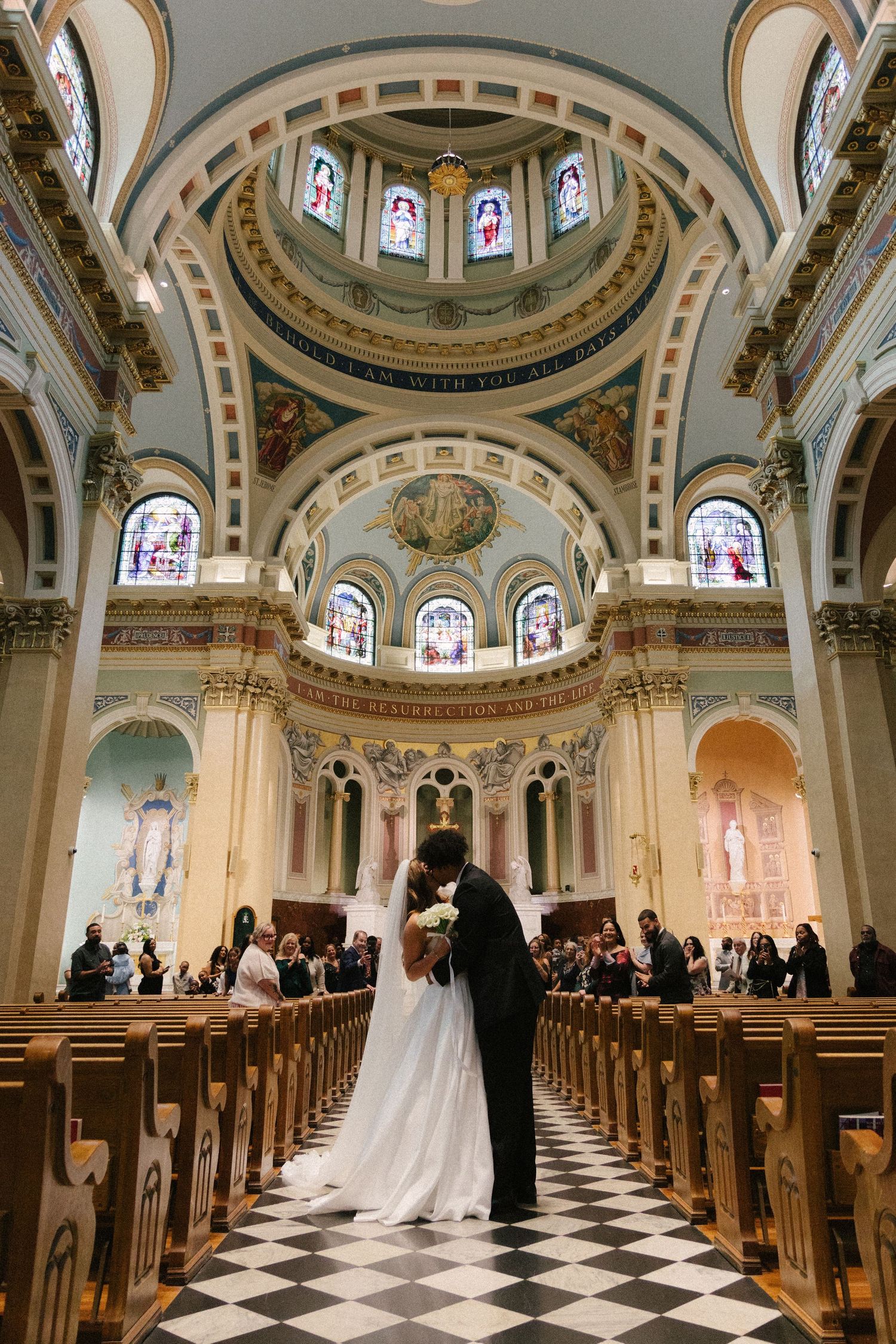 Wedding ceremony in an ornate cathedral with checkered floors and beautiful domed ceiling with stained glass windows.