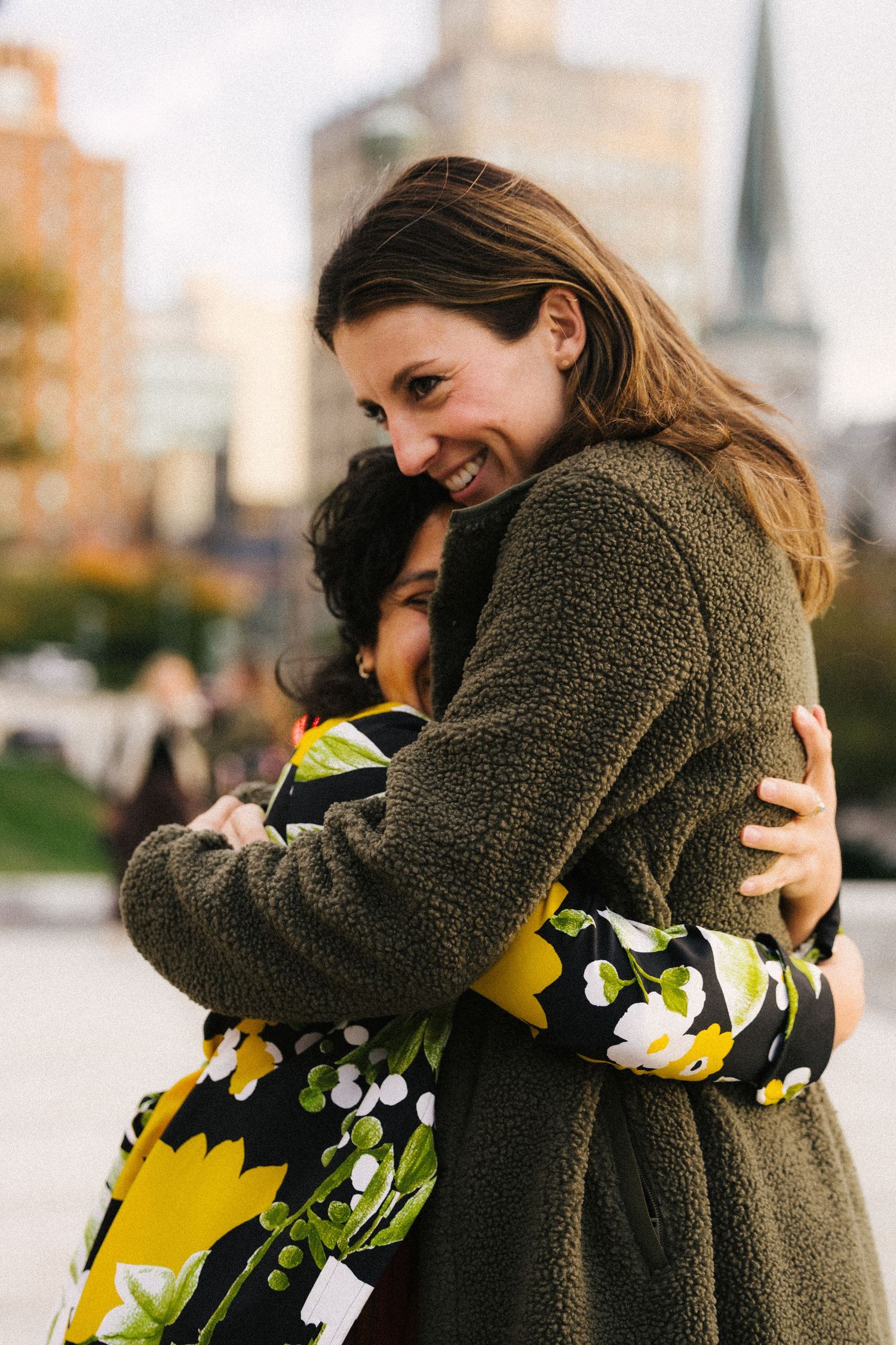 Two people share a warm embrace outdoors, one wearing a cozy olive green teddy coat and the other in a floral print dress.