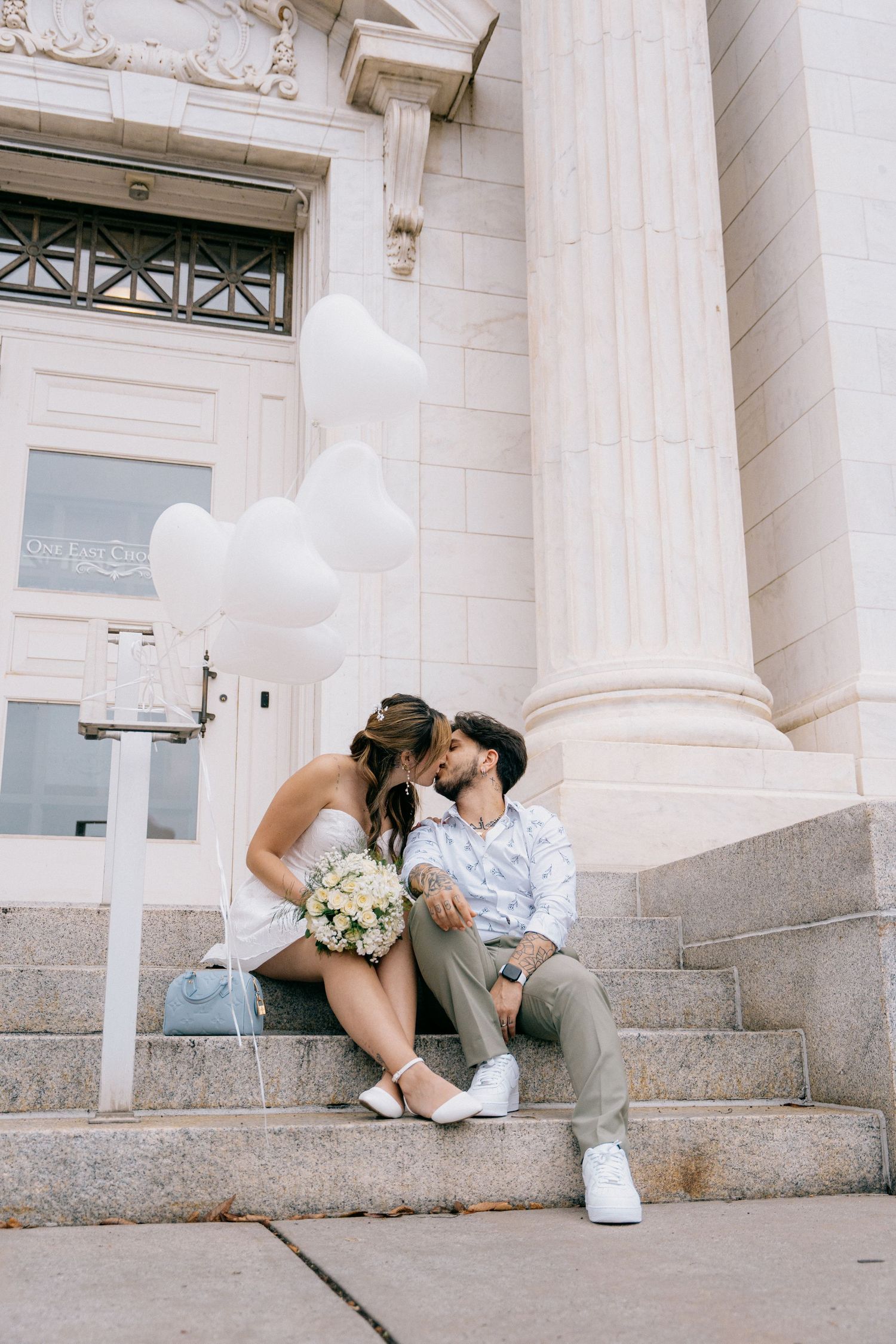 A couple shares a romantic moment sitting on stone steps outside a white columned building.