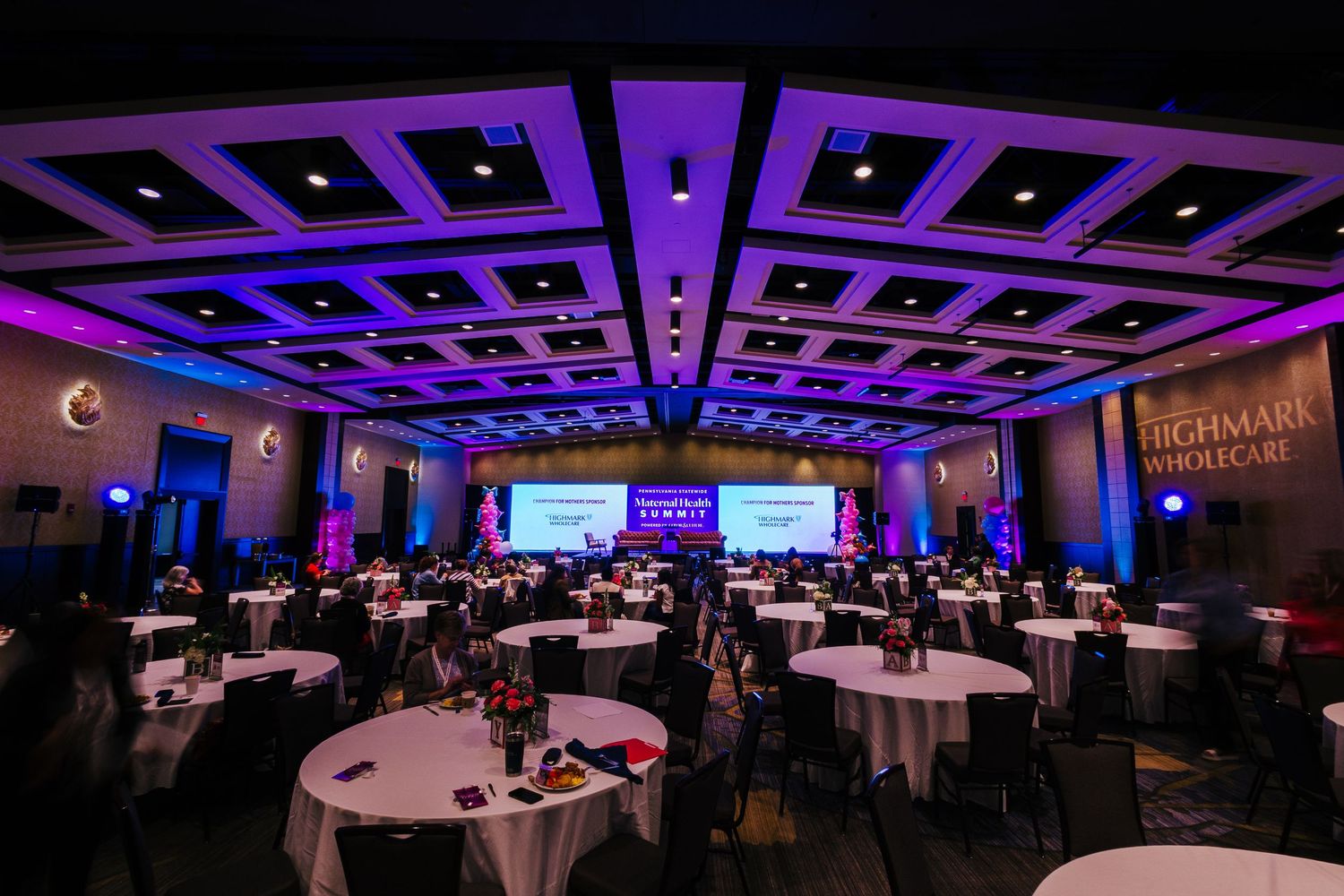 A panoramic view of a dimly lit ballroom with purple accent lighting and round tables set up for an event.