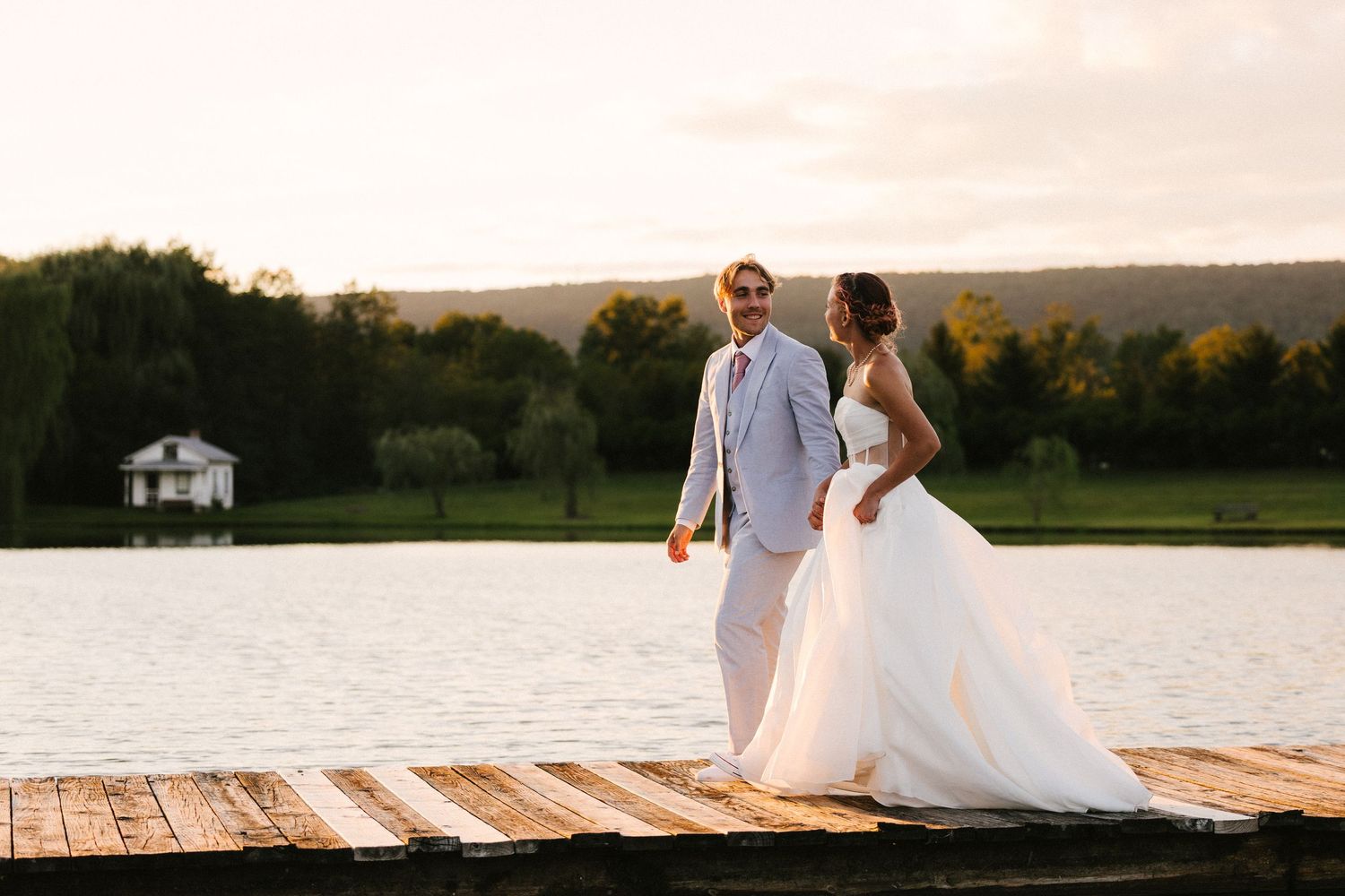 A couple in wedding attire poses on a wooden dock at sunset near a serene lake.