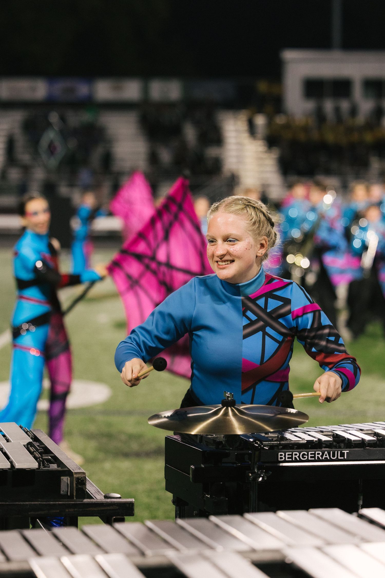 Smiling marching band performer playing Bergerault marimba in blue geometric uniform during colorful nighttime field show.
