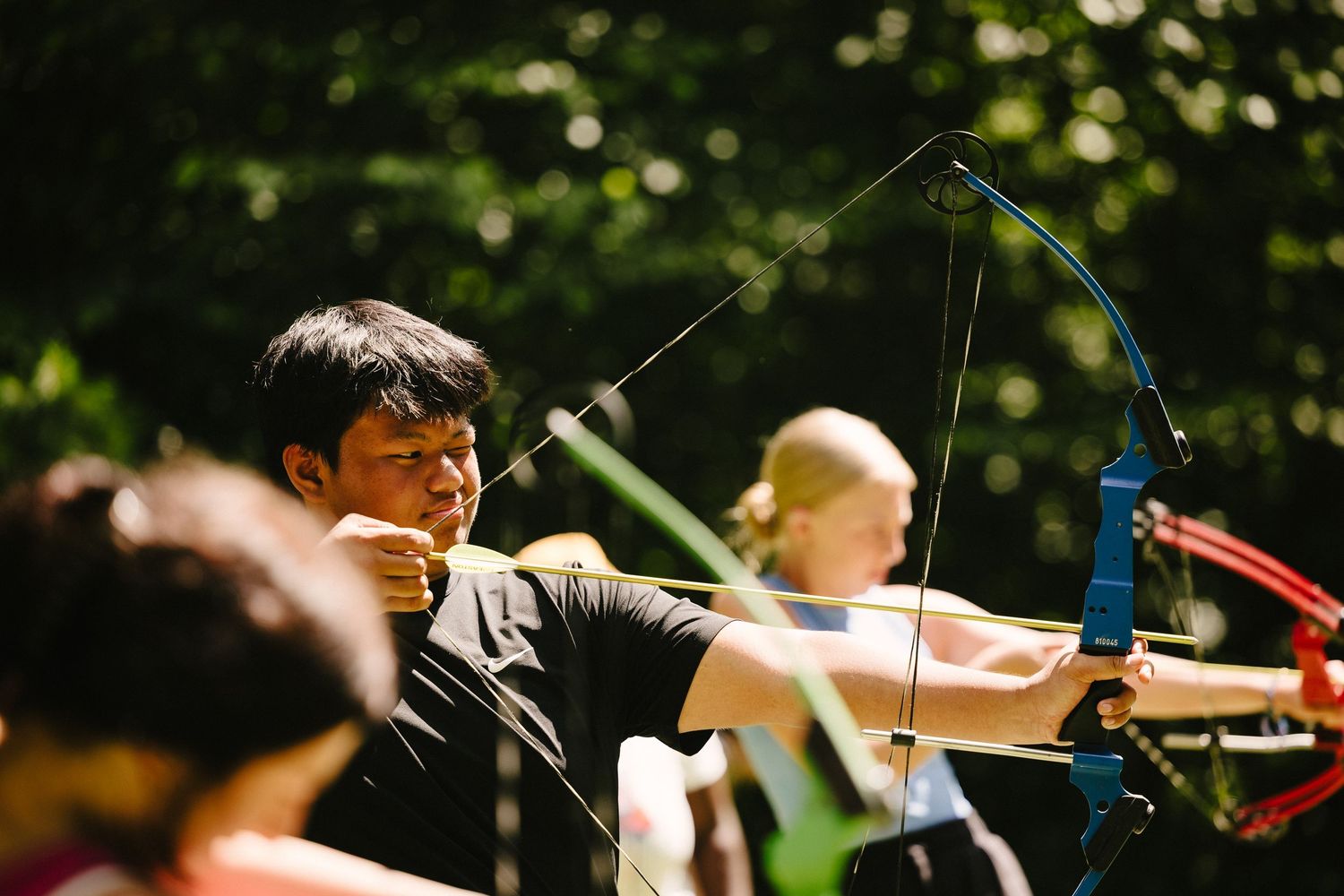 Archers practicing with bows at an outdoor archery range in a wooded setting during summer.