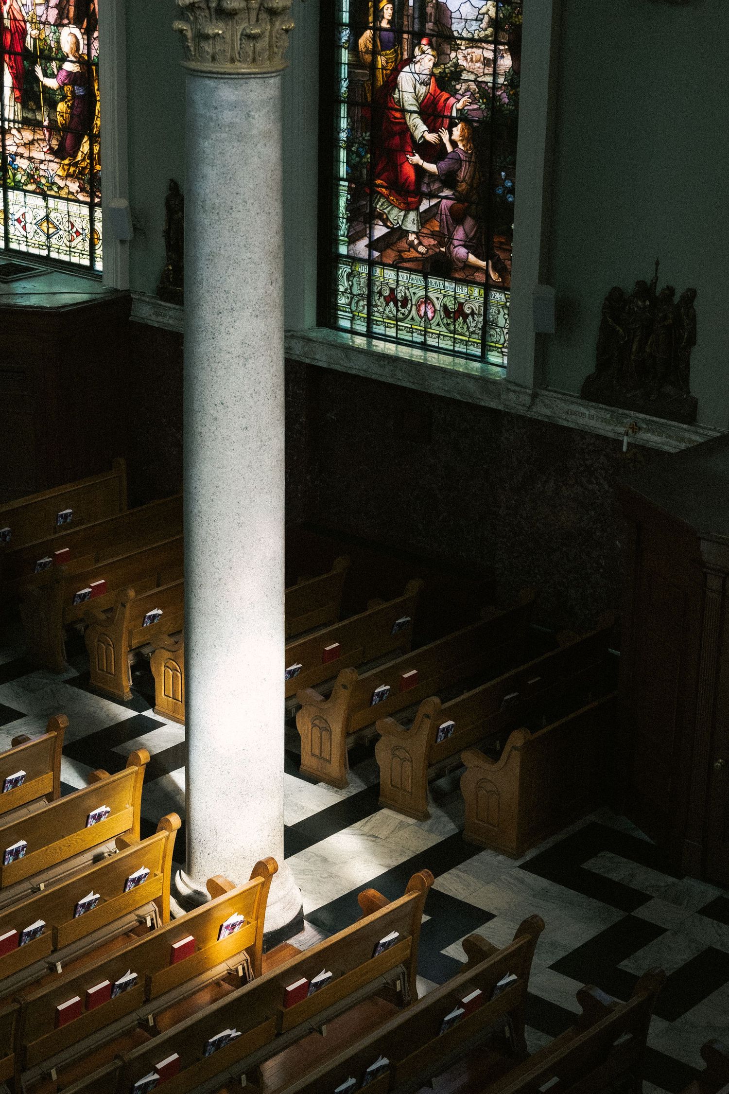 Sunlight streams through stained glass windows onto wooden church pews beside a white stone column in a cathedral interior.