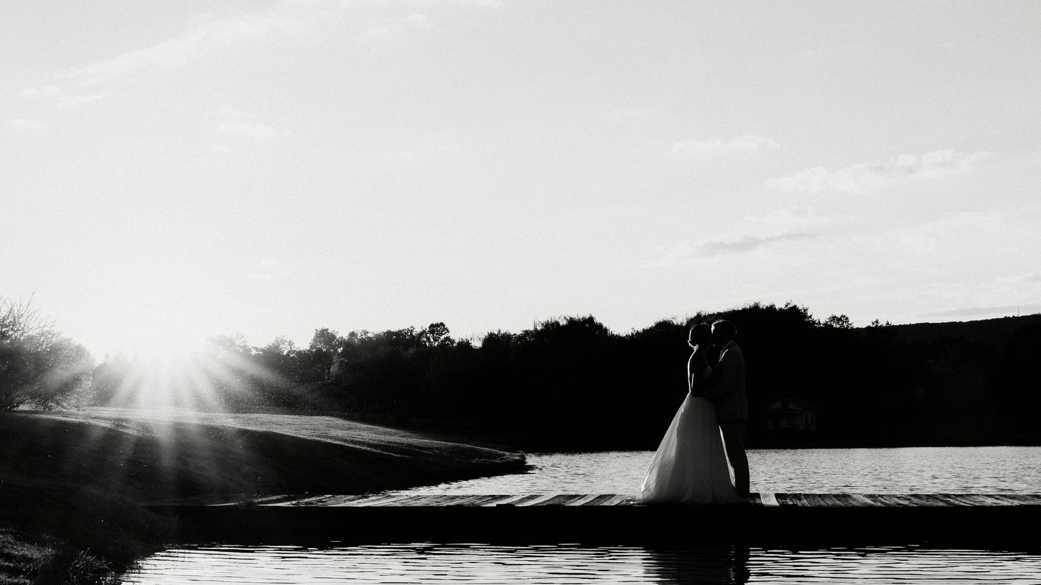 A panoramic black and white landscape photograph shows silhouetted trees against a cloudy sky with sunlight breaking through.