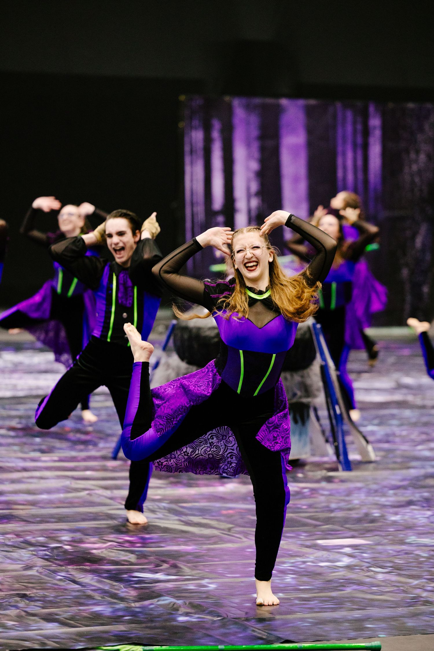 Indoor color guard performers dance in purple costumes during winter guard show.
