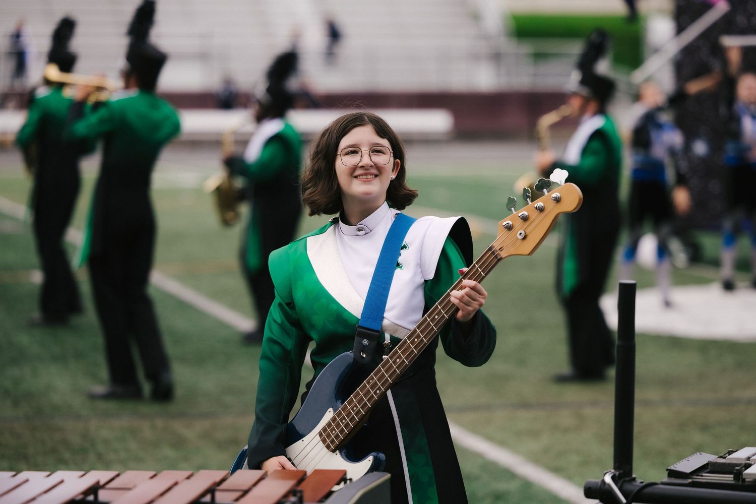 Smiling young woman playing bass guitar in green marching band uniform on football field during performance.