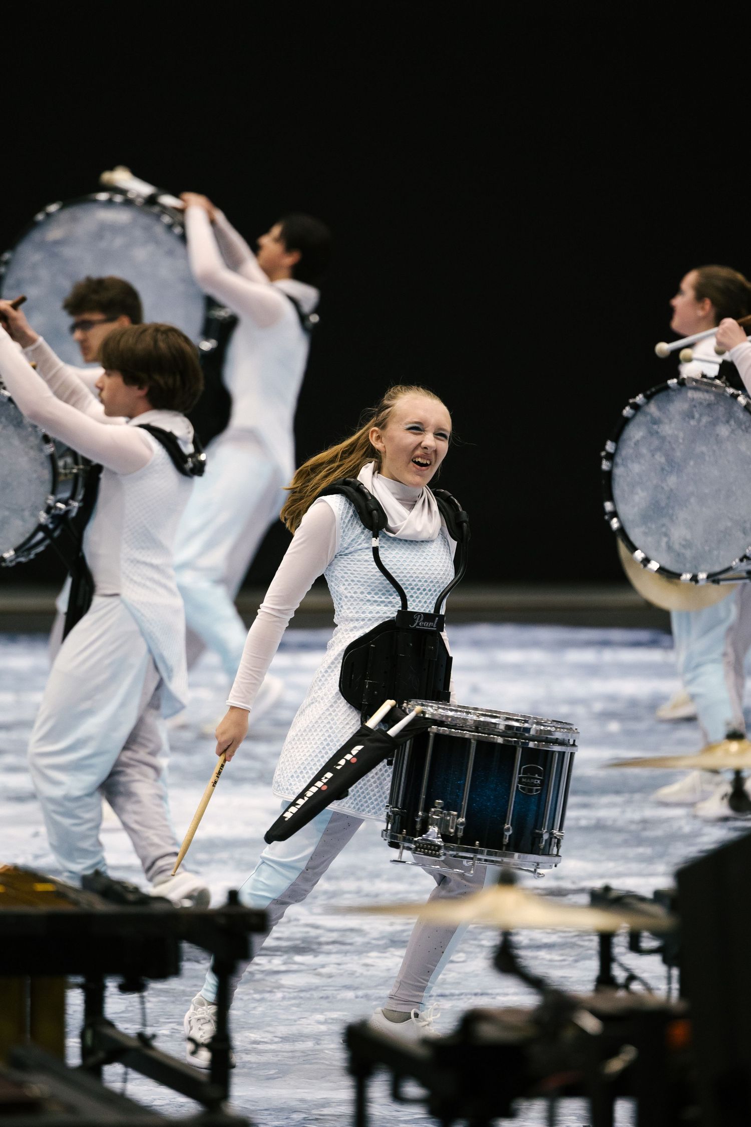Young female drummer smiles while performing with a marching percussion ensemble in white and black uniforms indoors.
