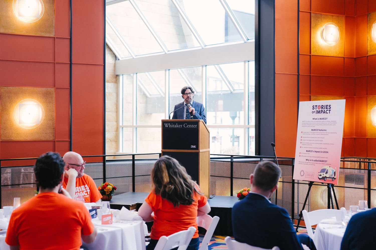 A speaker presents at a podium in front of large windows while attendees in red shirts listen at tables during an event.