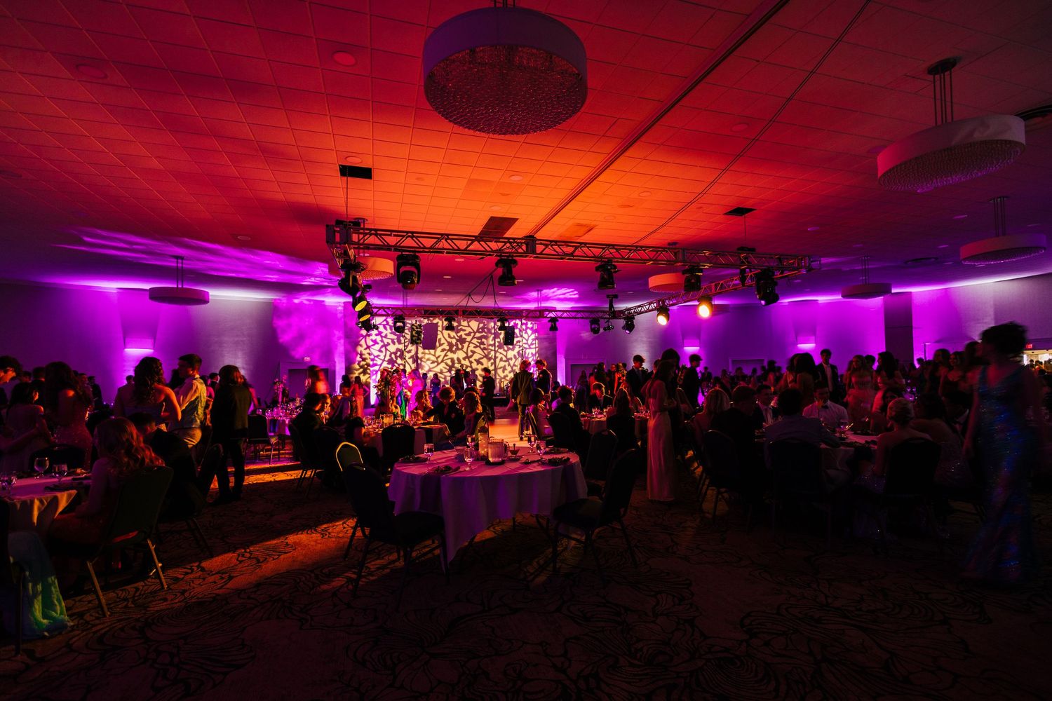 A dimly lit event venue with purple uplighting and a crowd gathered around tables during an evening celebration.