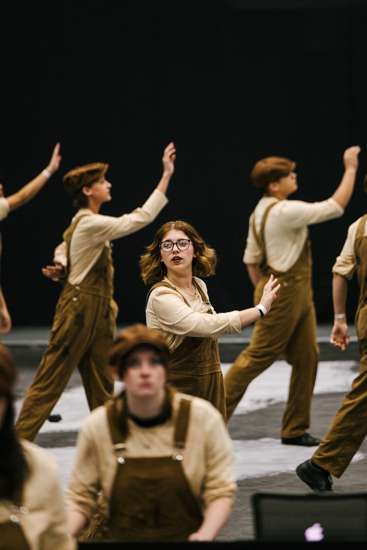 Performers in brown pants and beige shirts dance in formation on a dark stage during a theatrical production.