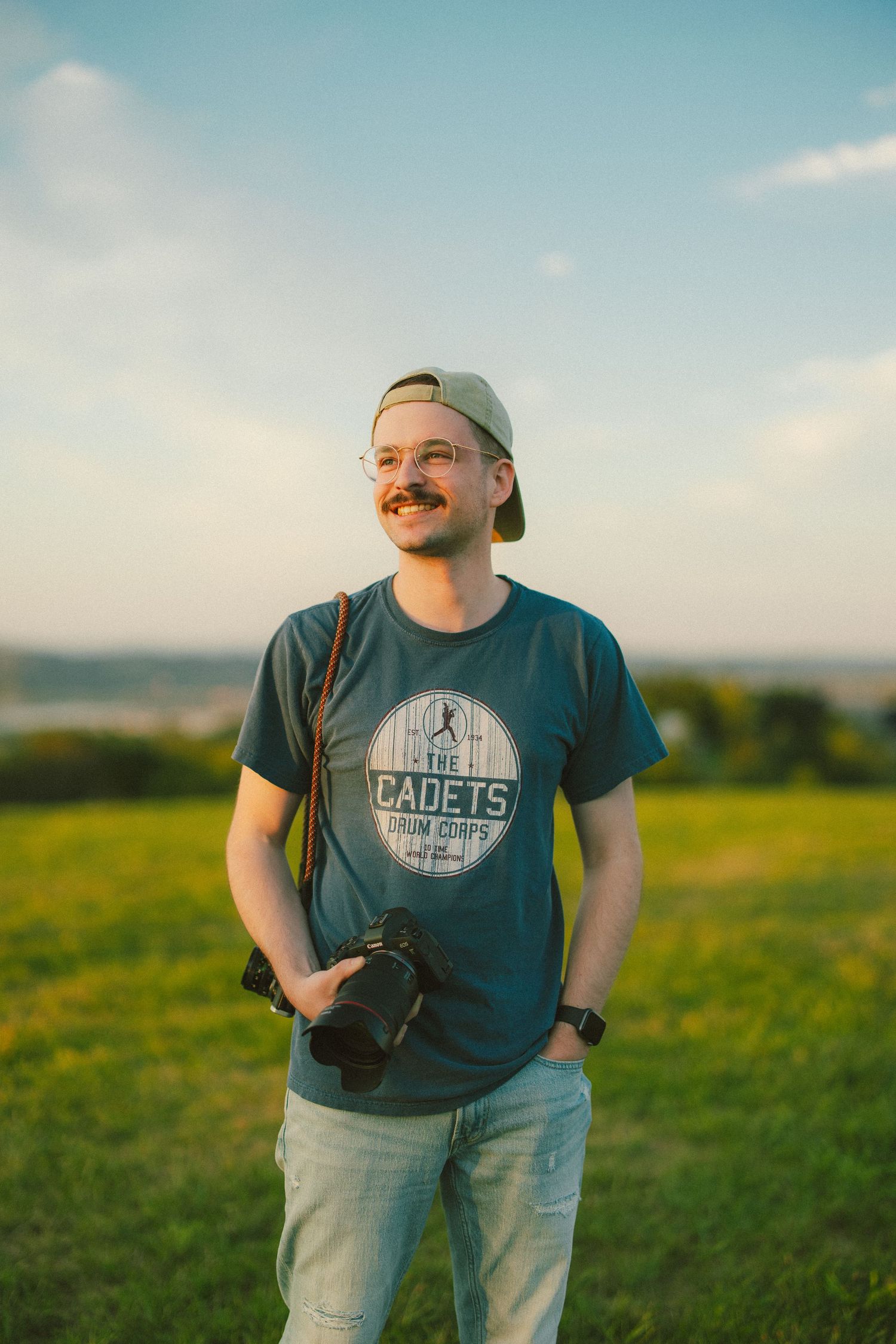 Person wearing a blue t-shirt with 'Cadets' logo stands in a sunlit grassy field during golden hour.