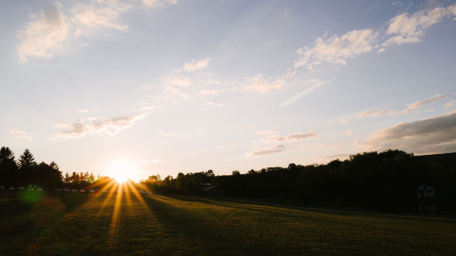 Panoramic sunset landscape with sunburst peeking through silhouetted trees against a cloudy orange and blue sky.