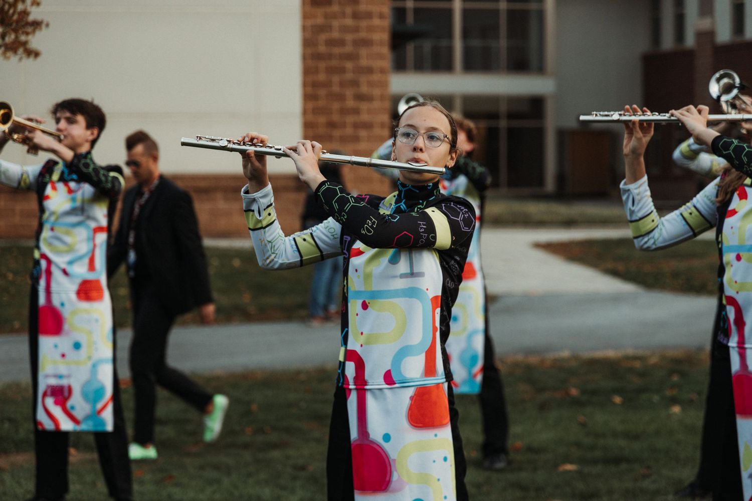 Marching band members practice their routine outdoors in front of a brick building.