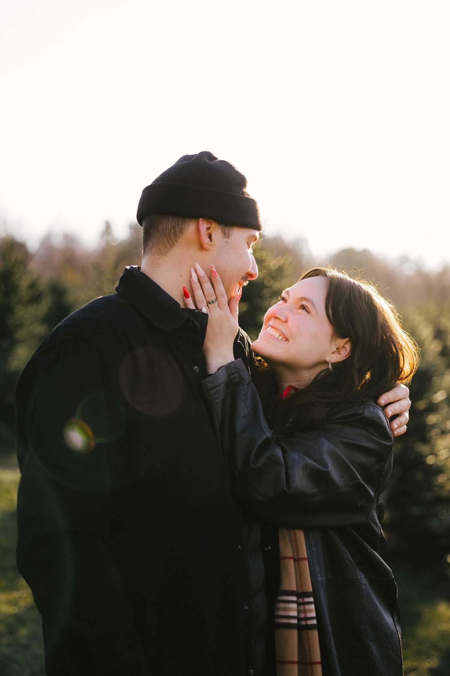A romantic couple in dark coats embrace and share intimate moments against a backlit natural setting at sunset.