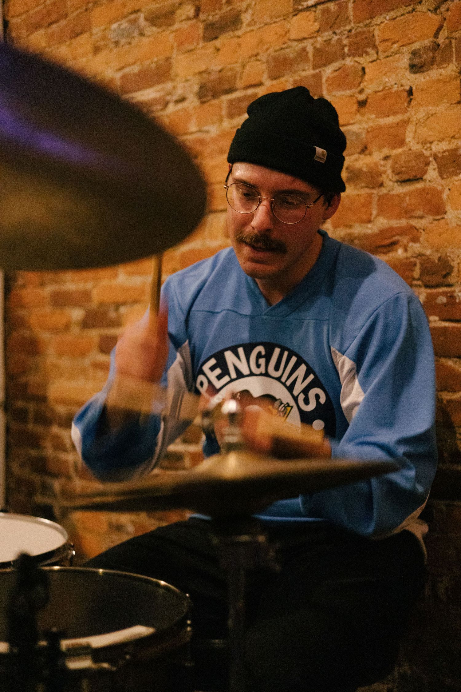 A drummer wearing glasses and a blue shirt performs behind a drum kit against an exposed brick wall.