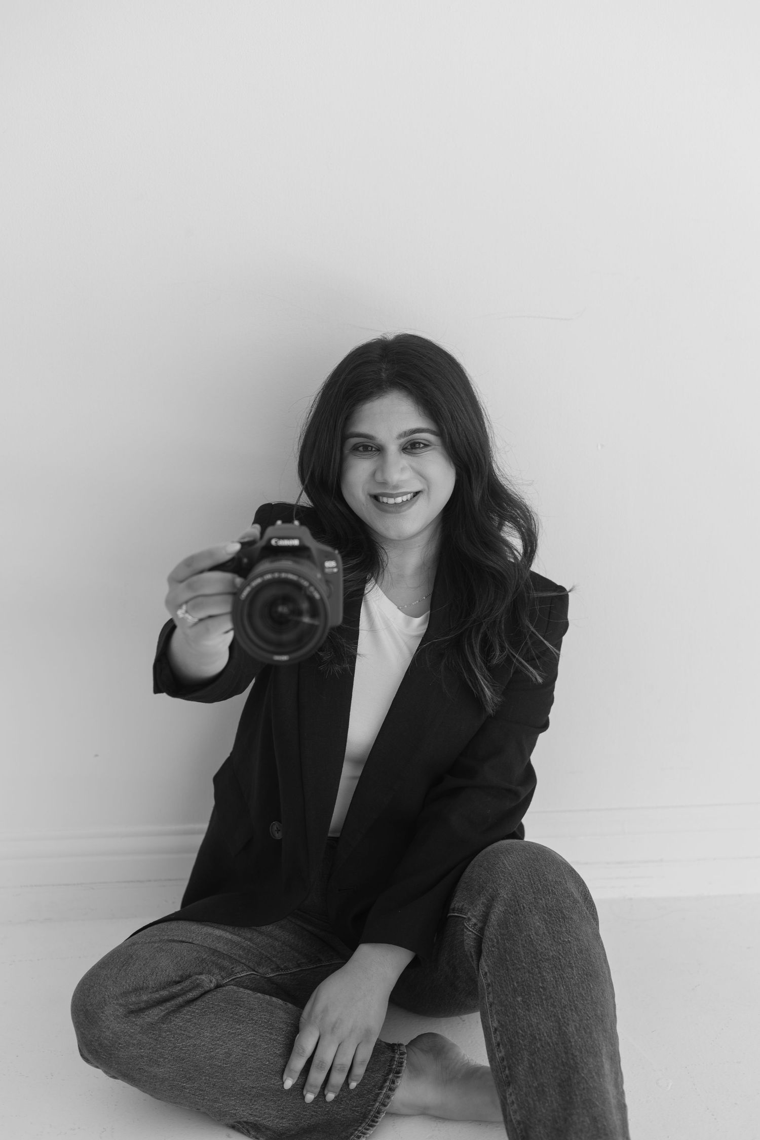 Black and white portrait of photographer sitting with camera against white wall.