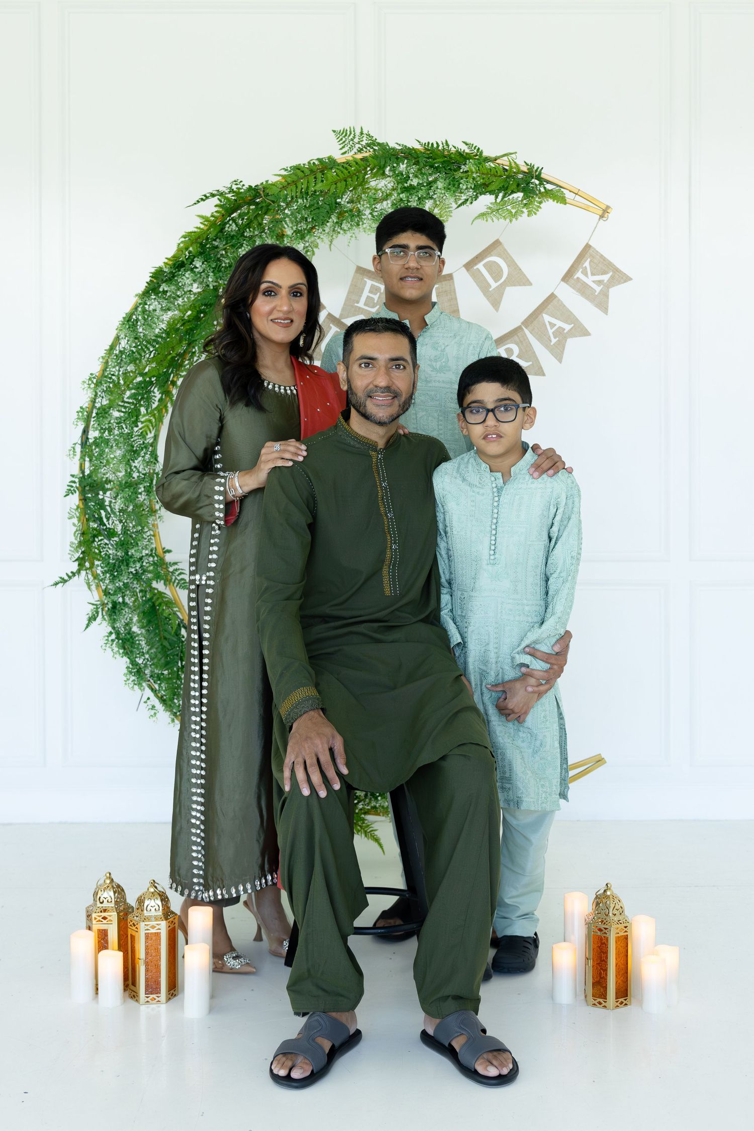 A family poses together in traditional Middle Eastern attire against a white backdrop with decorative greenery and lanterns.