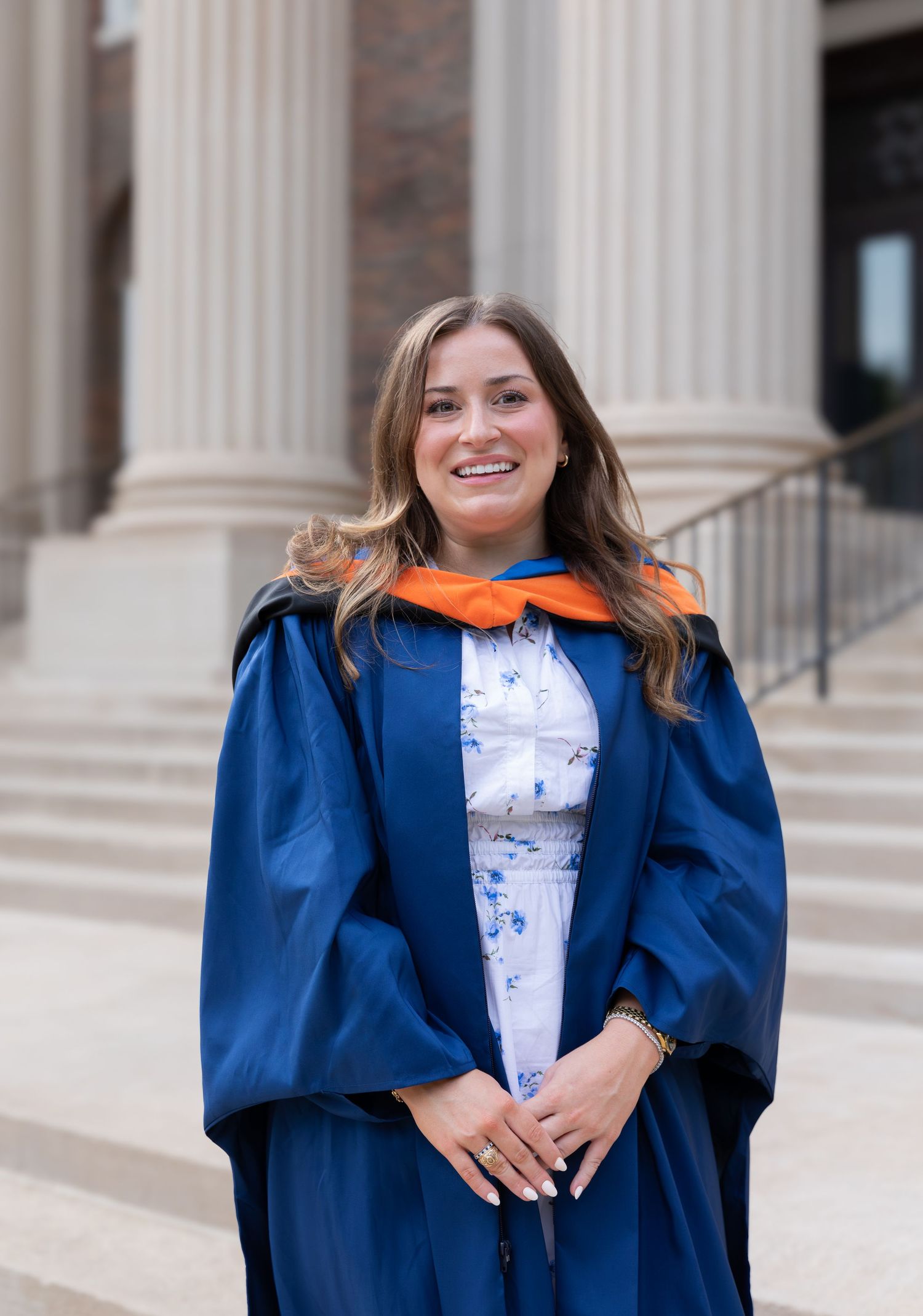 A graduate in blue academic regalia and orange stole stands proudly on stone steps.