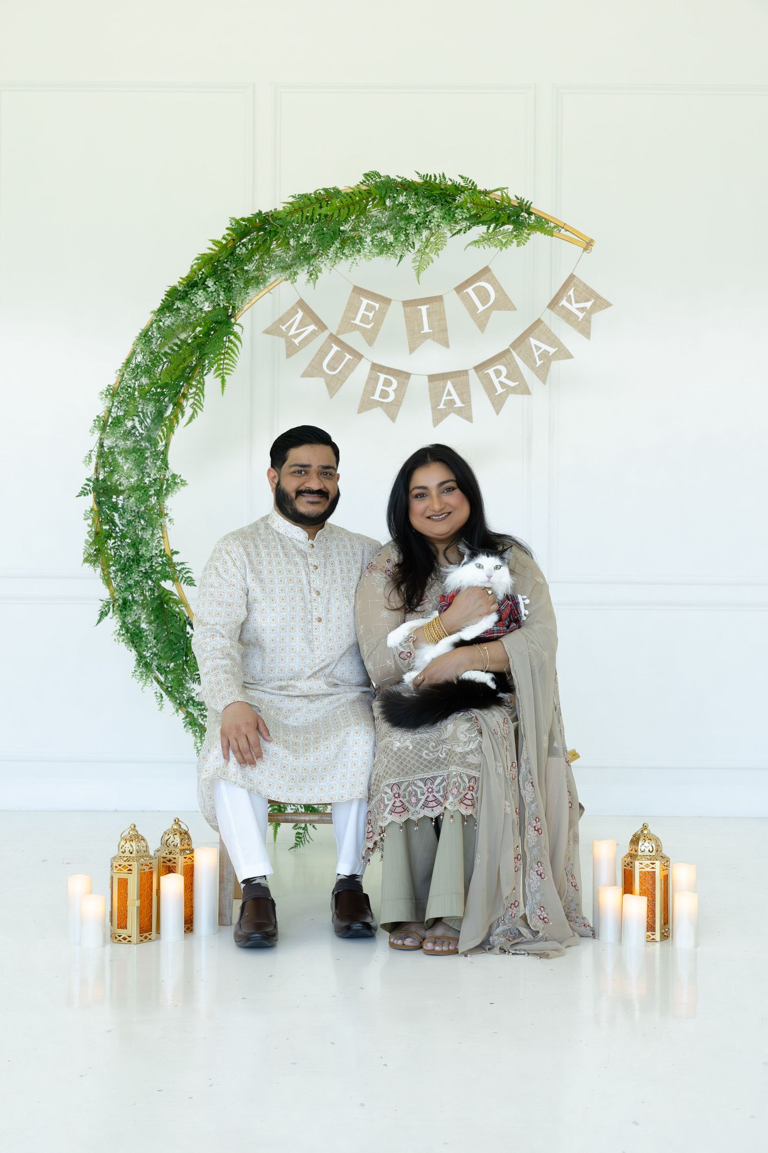 Couple in white and beige traditional outfits pose for Eid celebration photos with lanterns and greenery arch decor.