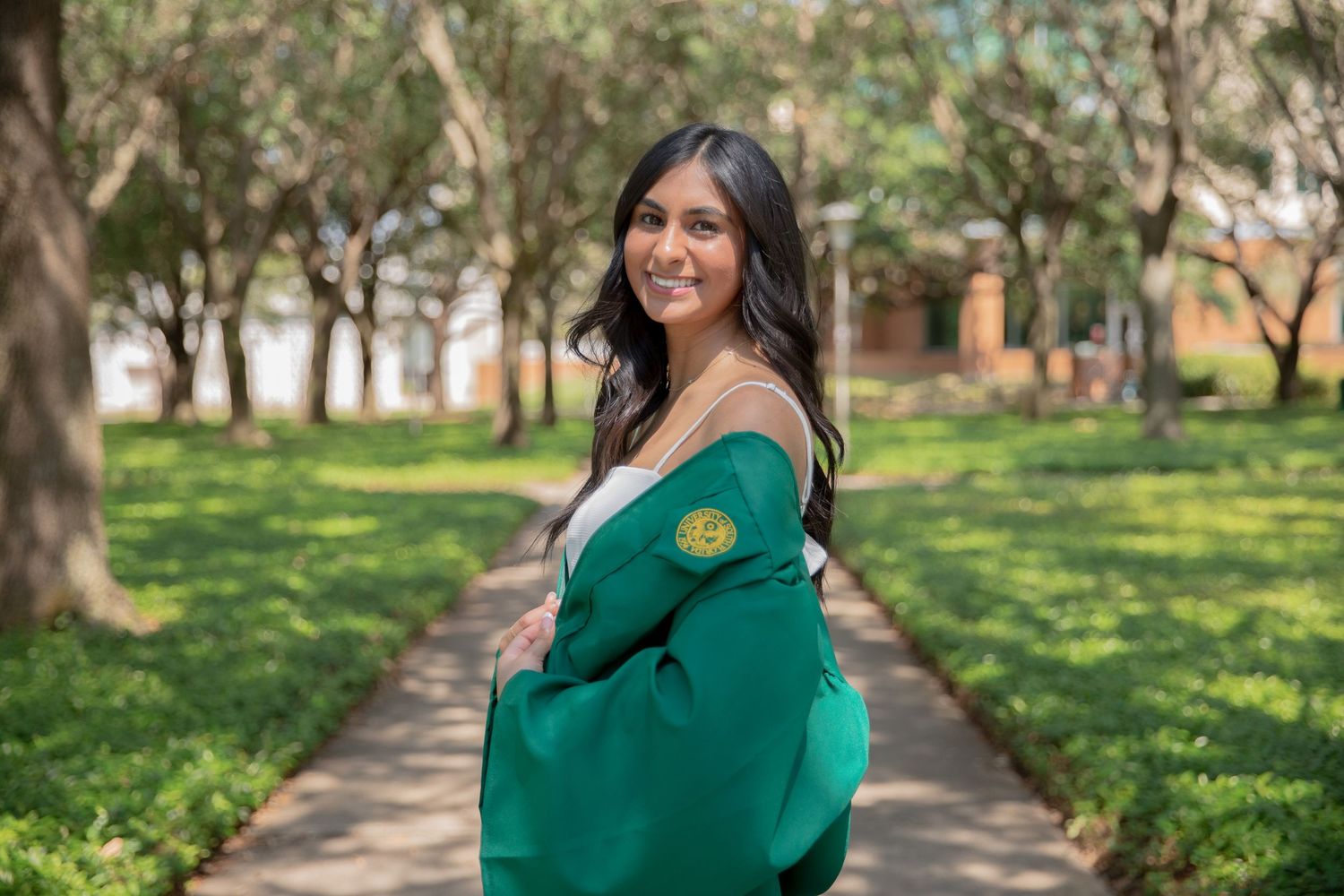 Graduate in green academic regalia smiling while posing on a tree-lined campus walkway.