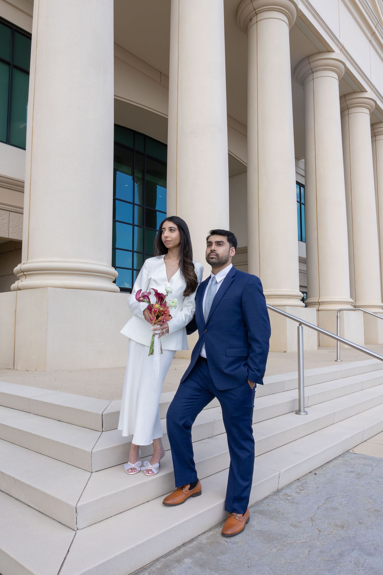 Couple in formal attire on building steps, woman in white suit holding bouquet, man in navy suit, elegant columns backdrop.