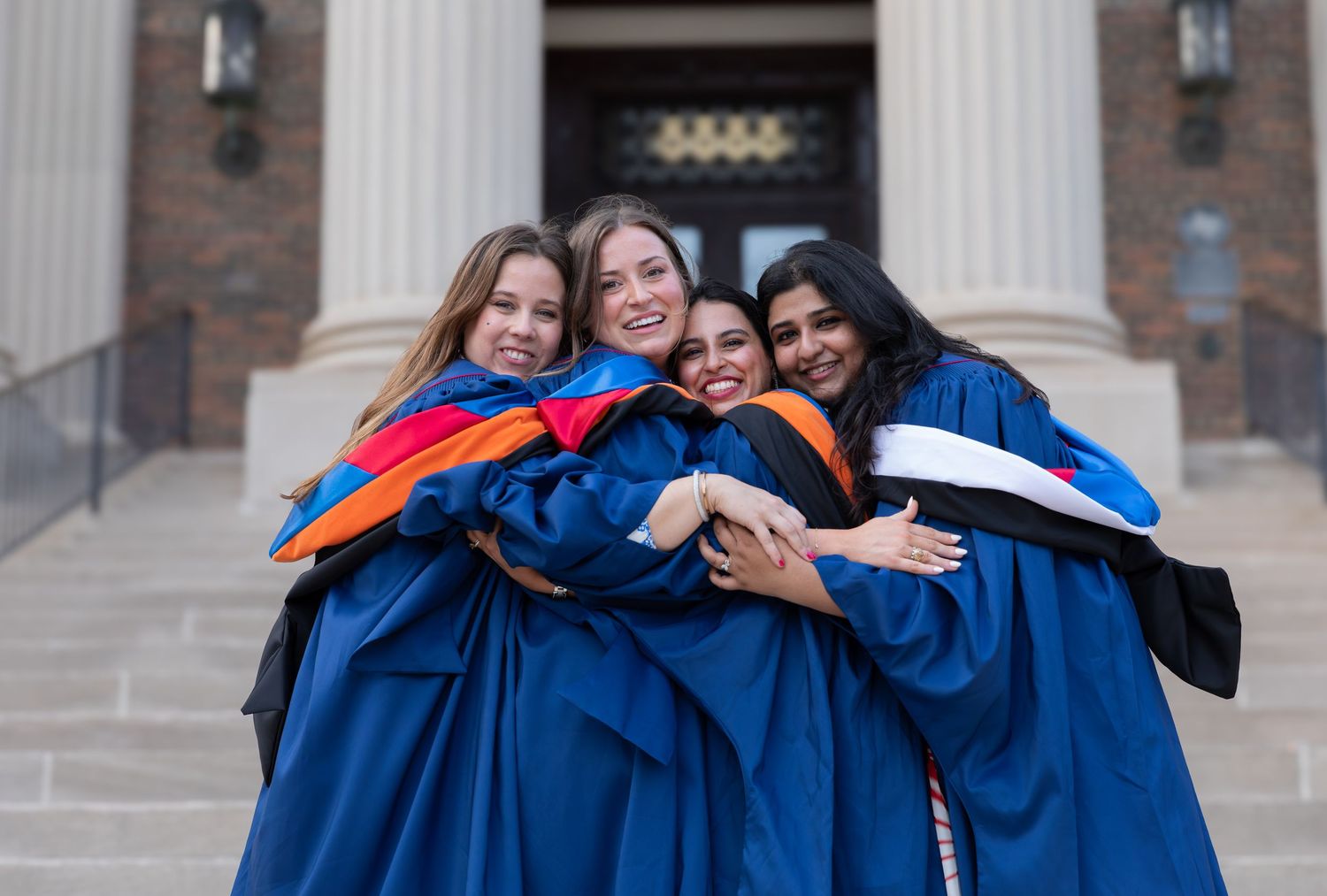 Graduates in blue caps and gowns celebrating with colorful stoles on campus steps.