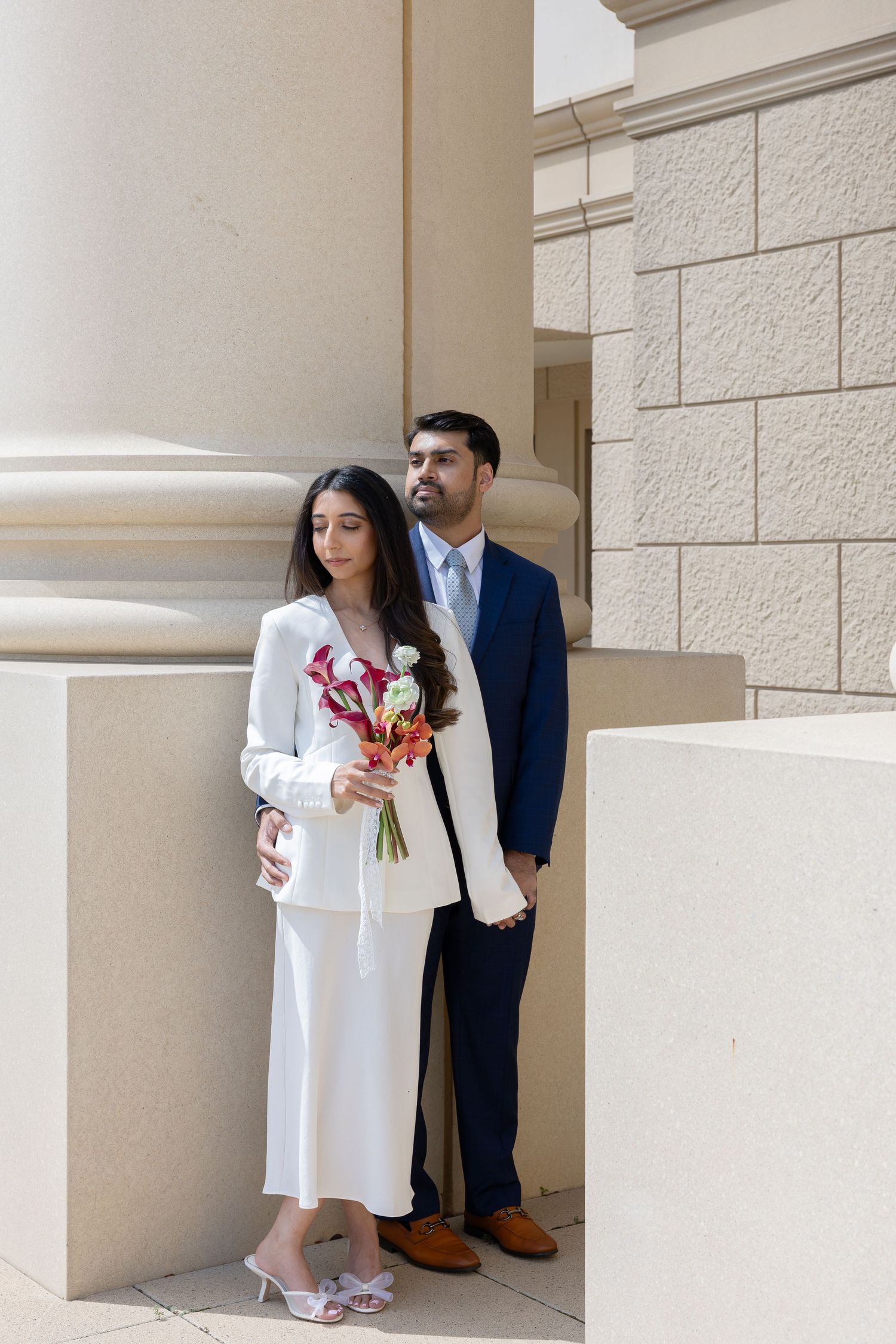 Bride in white suit holding bouquet stands with groom in navy suit by stone columns at elegant venue.