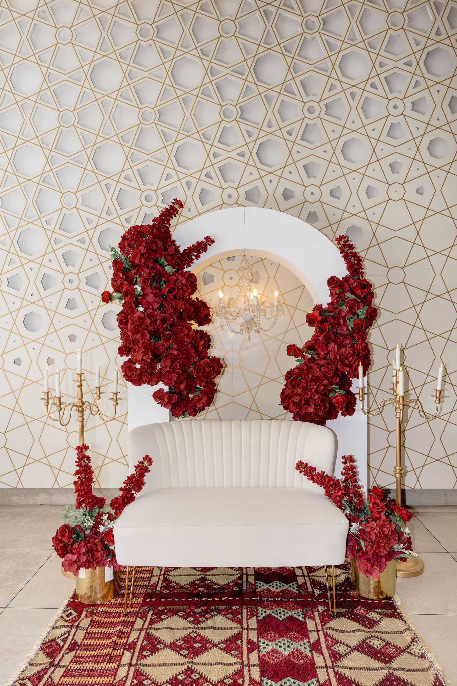 Elegant white wedding throne chair adorned with cascading red roses, gold candelabras, and patterned rug backdrop.