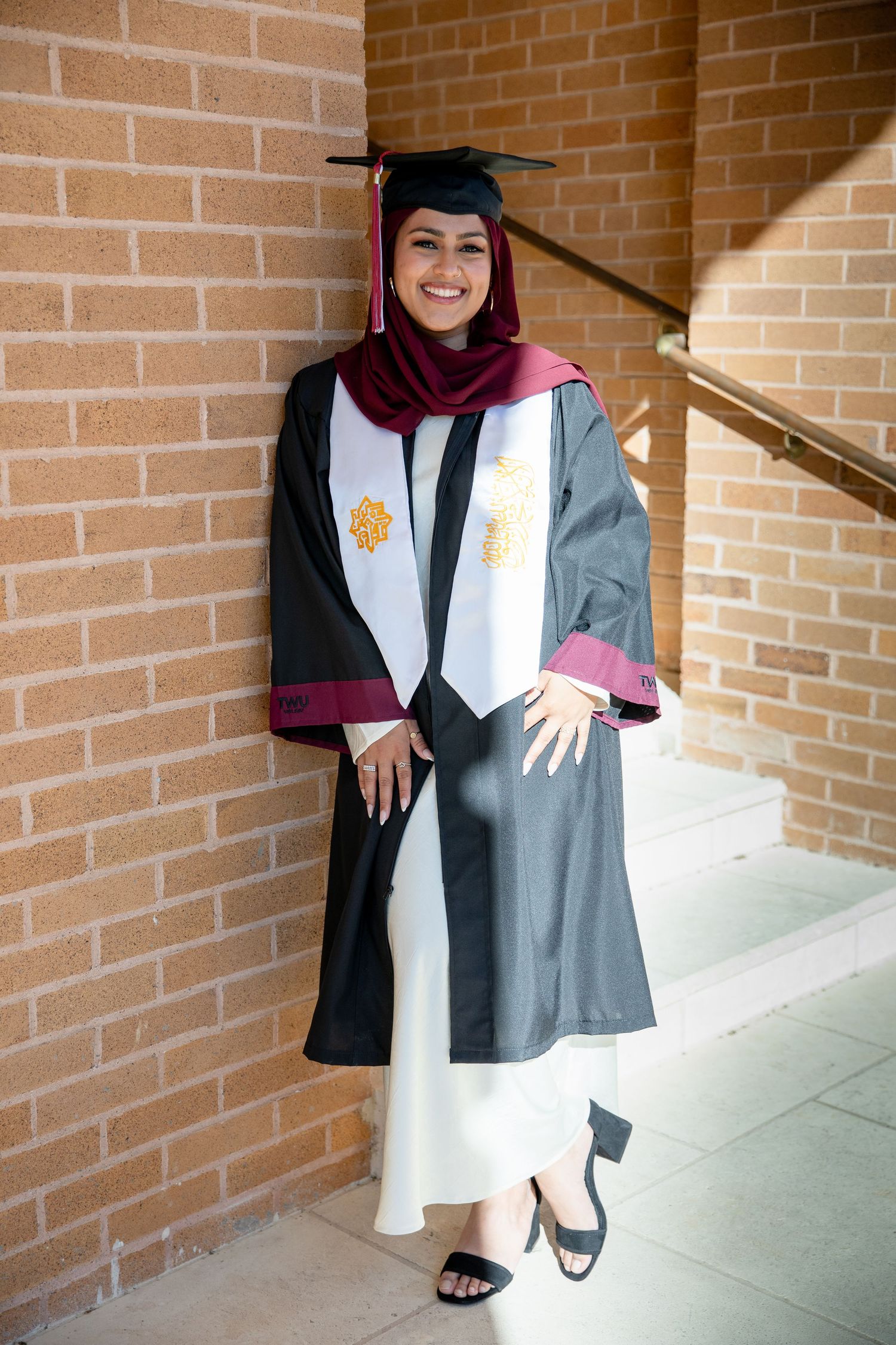 Smiling graduate in black gown, white stole, and burgundy hijab poses against brick wall.
