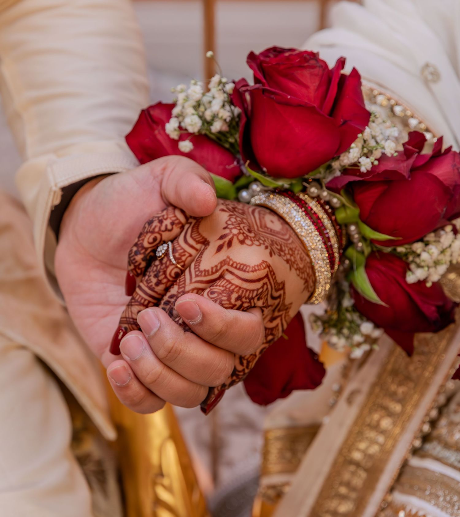 Hands adorned with intricate henna designs hold red roses with baby's breath flowers against a cream-colored background.