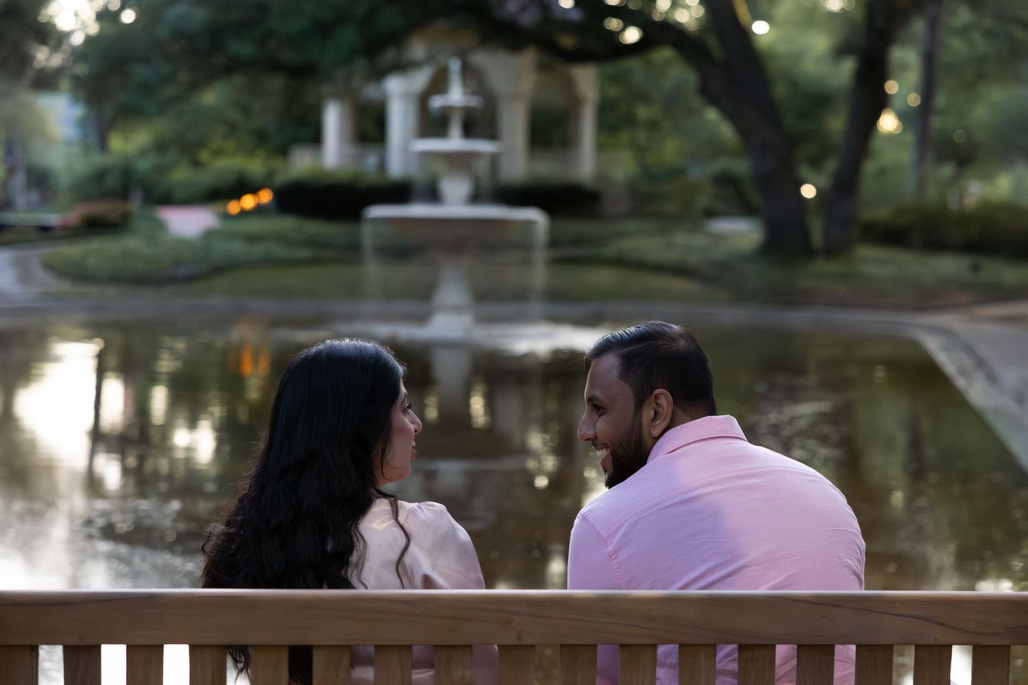 Couple sitting on a bench facing each other near a serene pond with a fountain in a lush park setting.