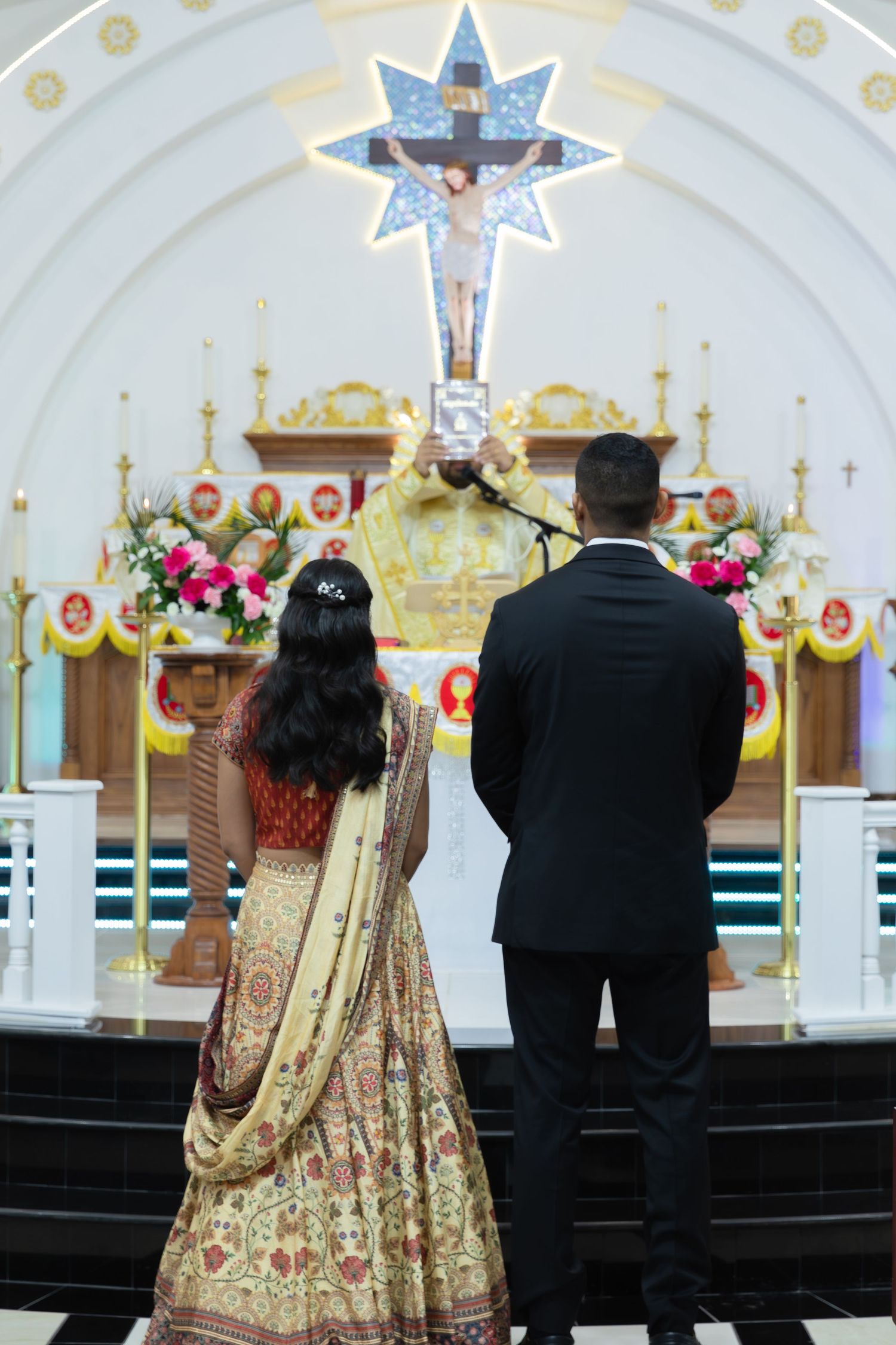 A couple in traditional wedding attire stands at the altar of an ornate church decorated with pink flowers and gold accents.