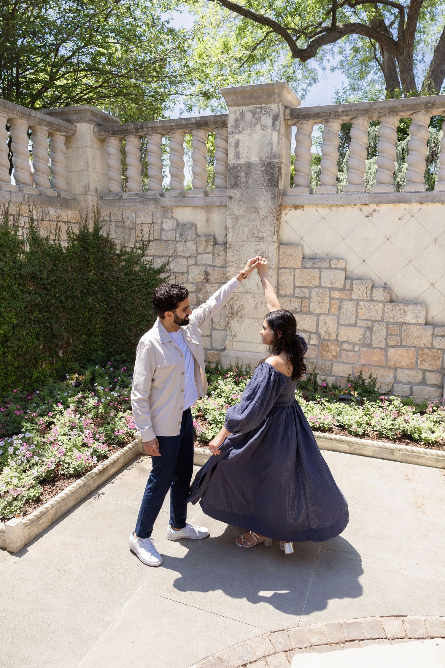Couple dancing outdoors in a garden, man in blazer spinning woman in flowing blue dress near stone balustrade.