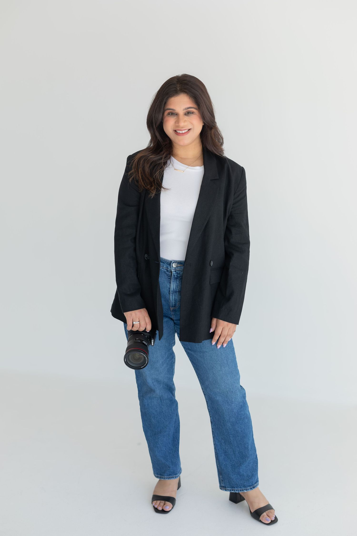 Creative professional in black blazer holding camera against white studio backdrop.