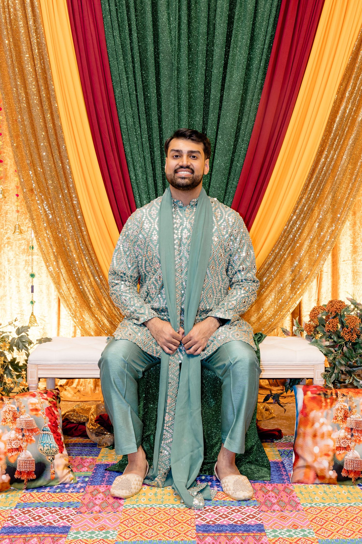 Man in elegant teal sherwani sits smiling before colorful draped backdrop at South Asian celebration.