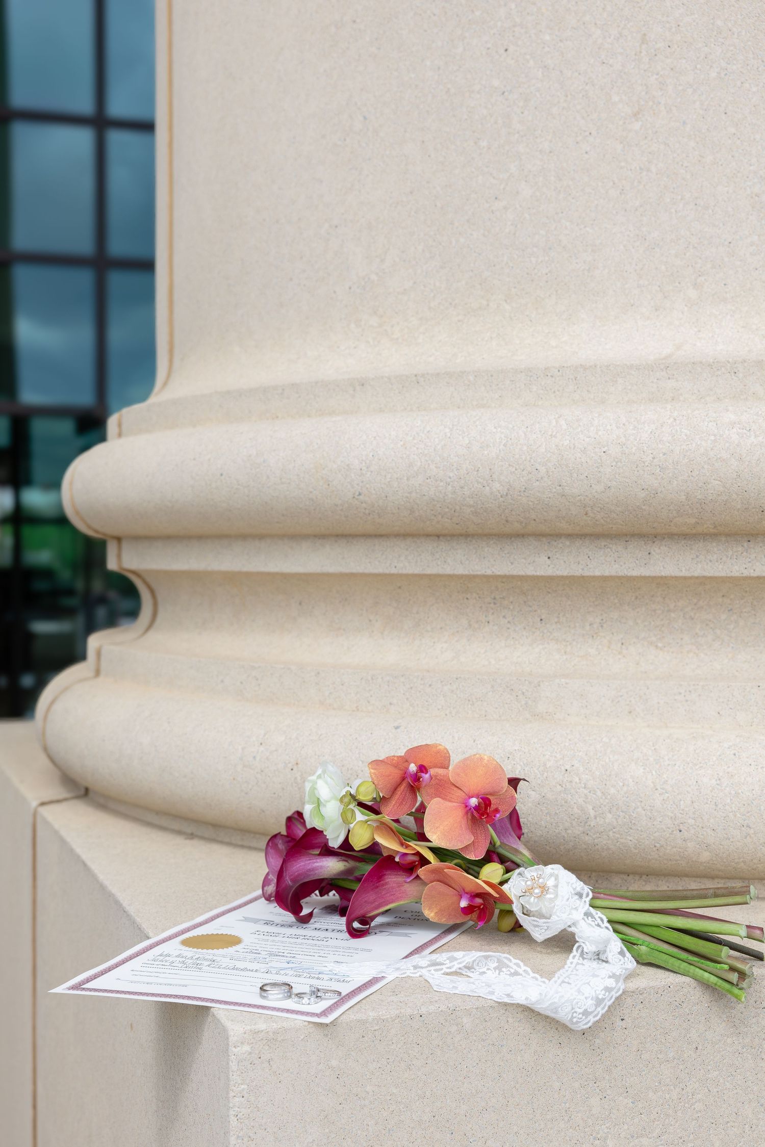 Colorful wedding bouquet with orchids and calla lilies resting beside a marriage certificate at a stone column base.