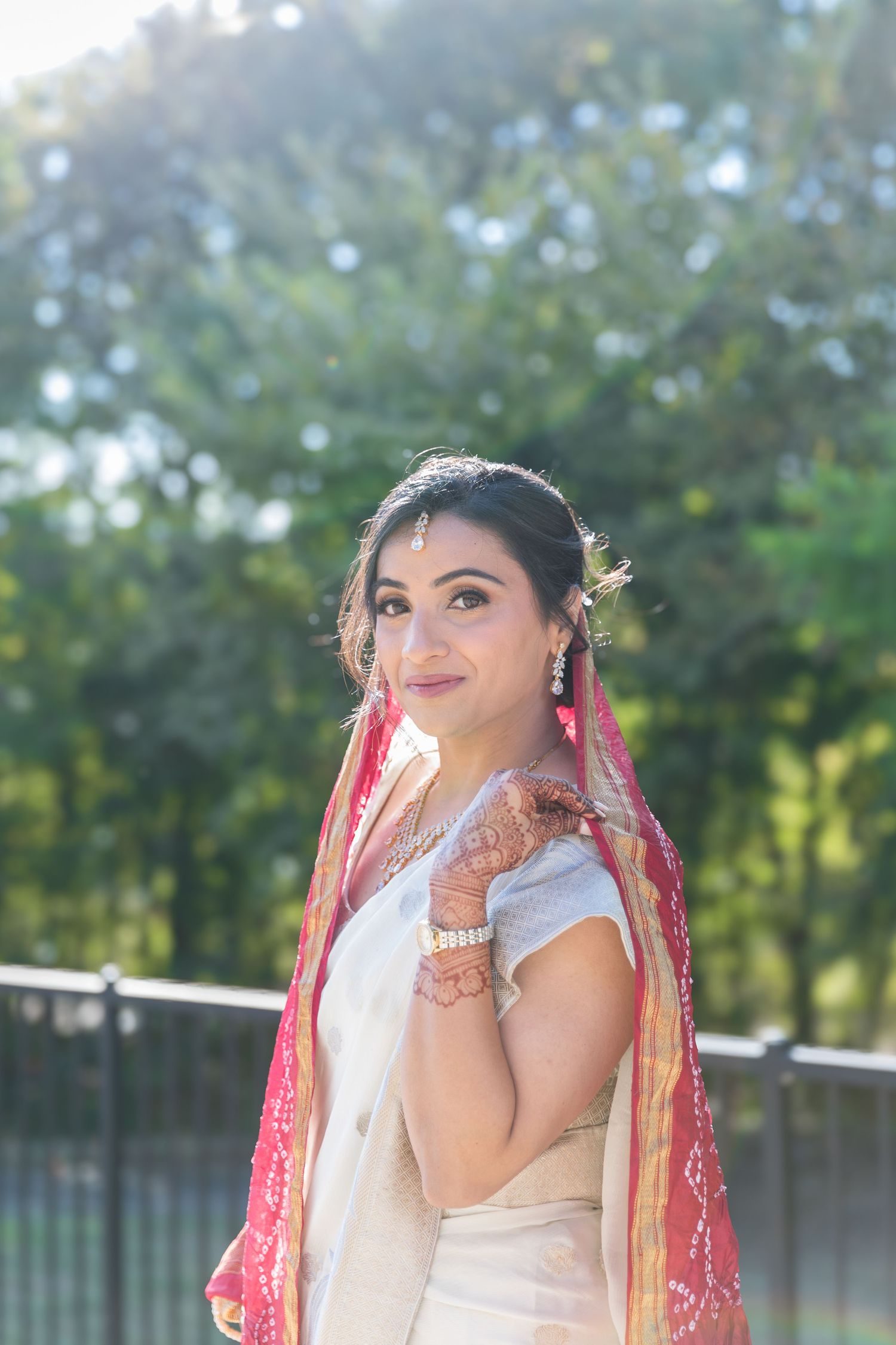 A bride in traditional South Asian wedding attire poses outdoors in a garden setting with natural lighting.