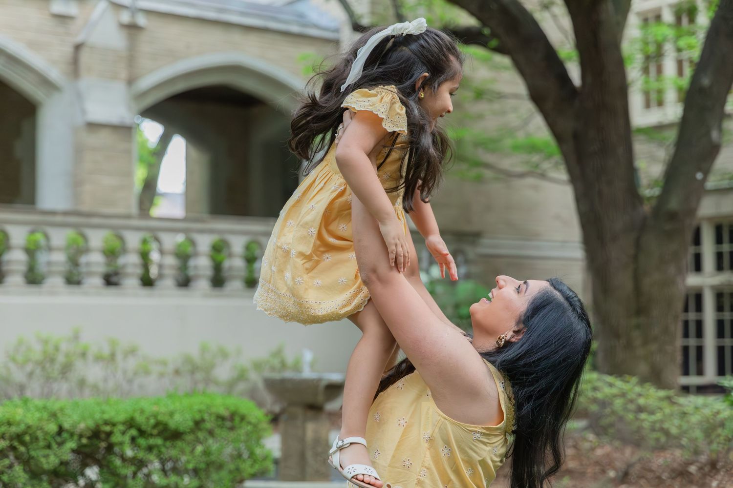 A mother in a yellow dress joyfully lifts her young daughter in a yellow dress outdoors near a stone building.