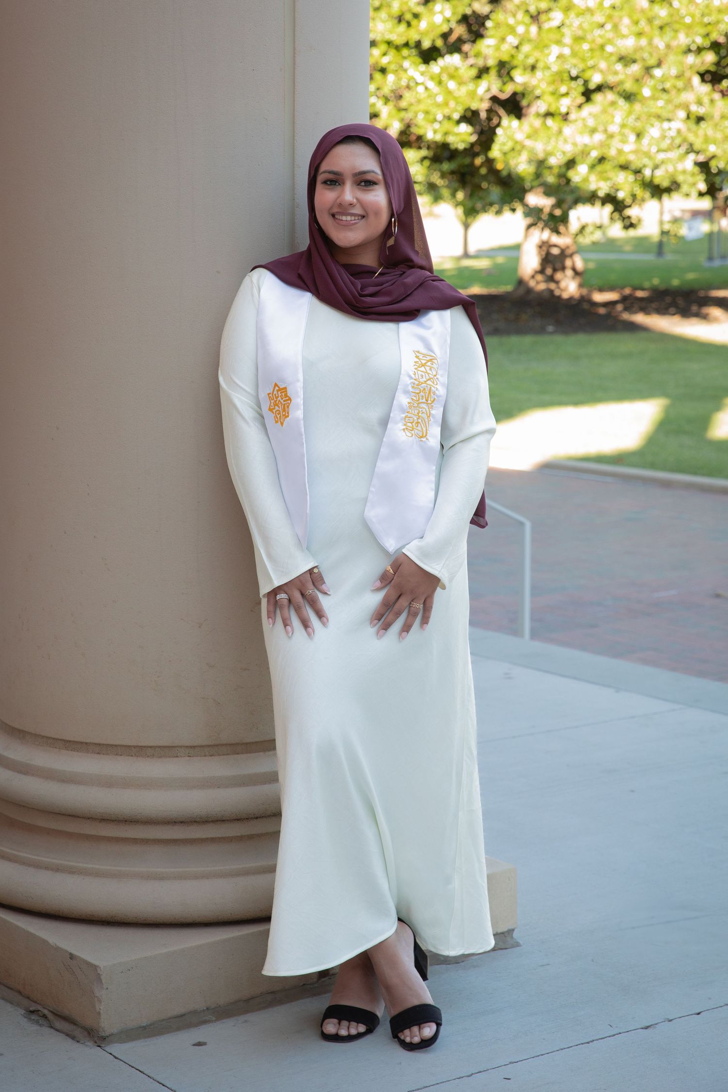 Woman wearing a white embroidered dress and burgundy hijab poses elegantly by a classical column.