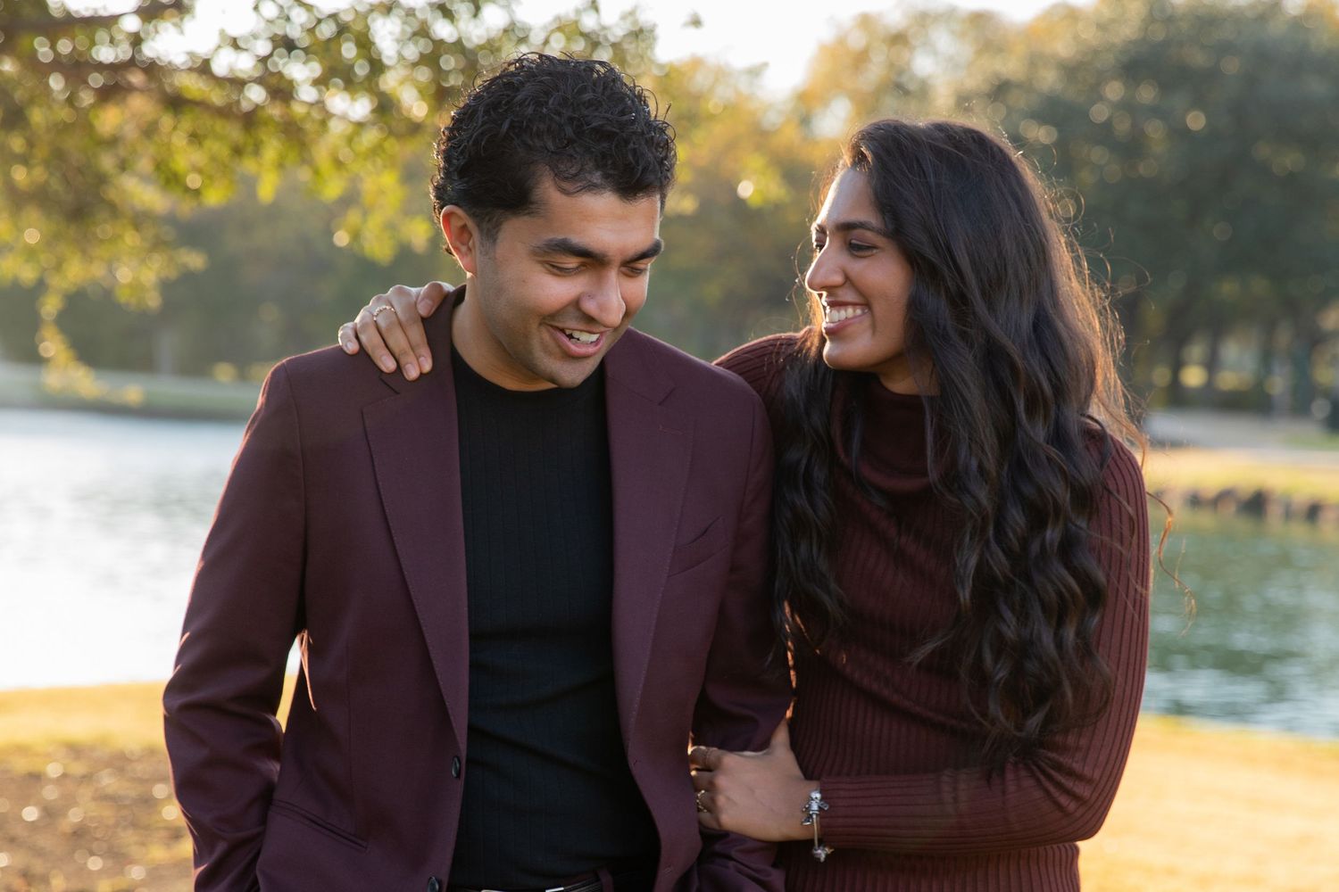 A couple in burgundy autumn attire sharing a joyful moment by a lakeside at sunset.