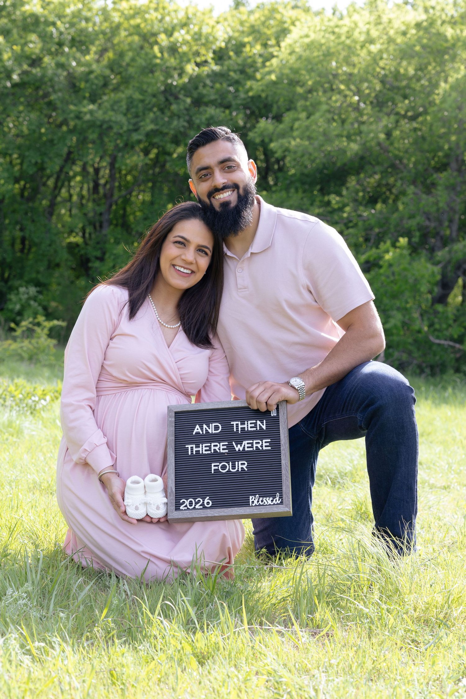 Couple poses outdoors holding pregnancy announcement sign reading 'And Then There Were Four 2026 Blessed' with baby shoes.