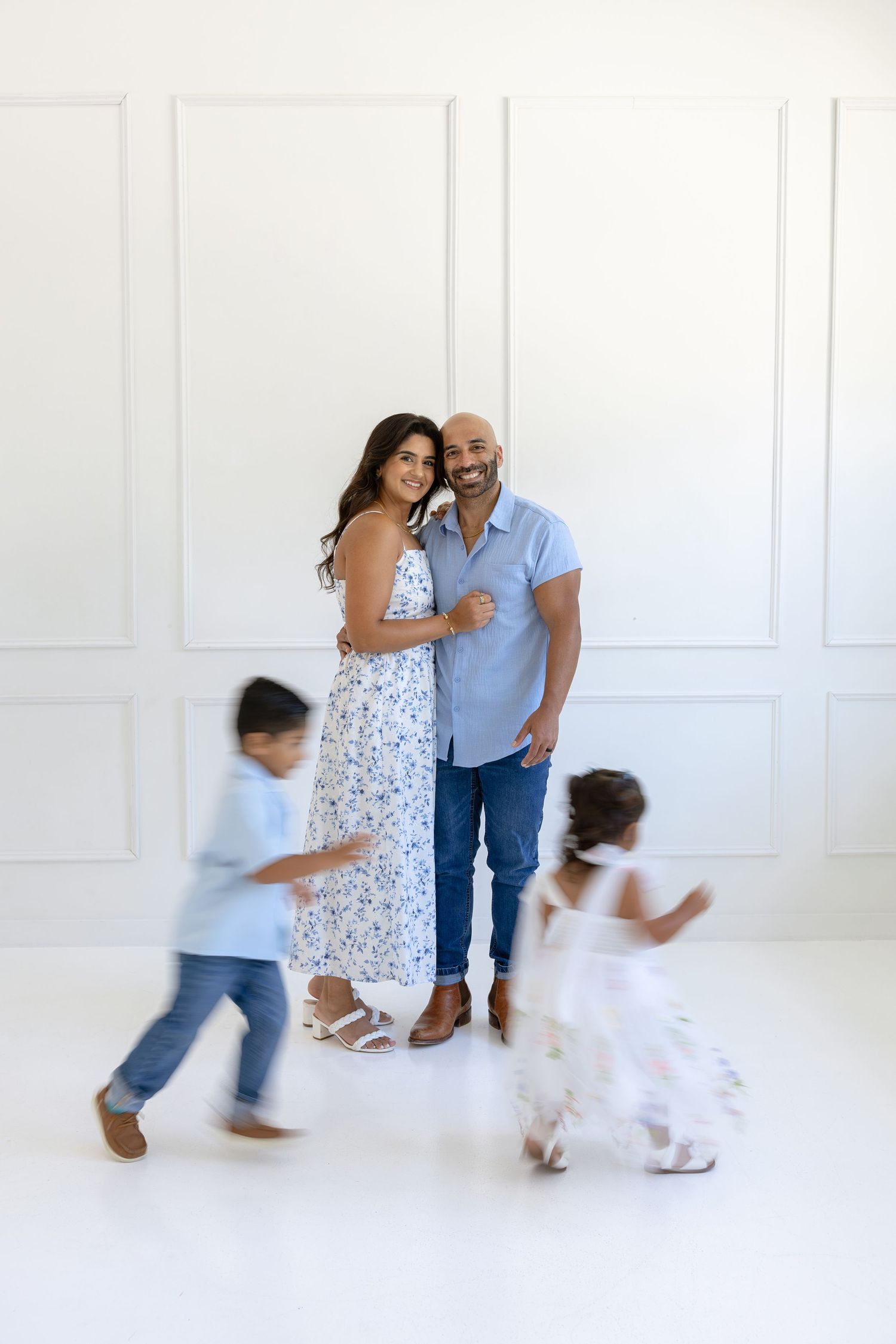 A smiling couple poses together while two young children run playfully in front of them in a bright white studio.