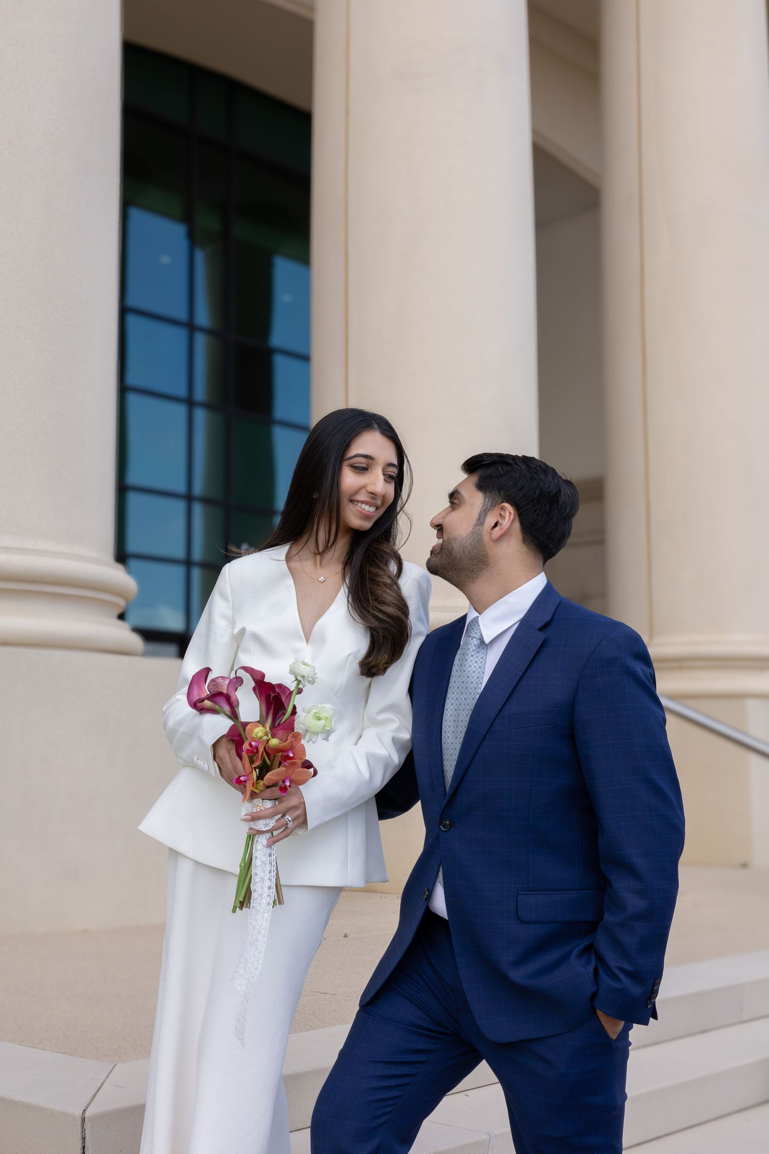 Couple in wedding attire, bride in white suit holding red bouquet, groom in navy suit, standing by elegant columns.