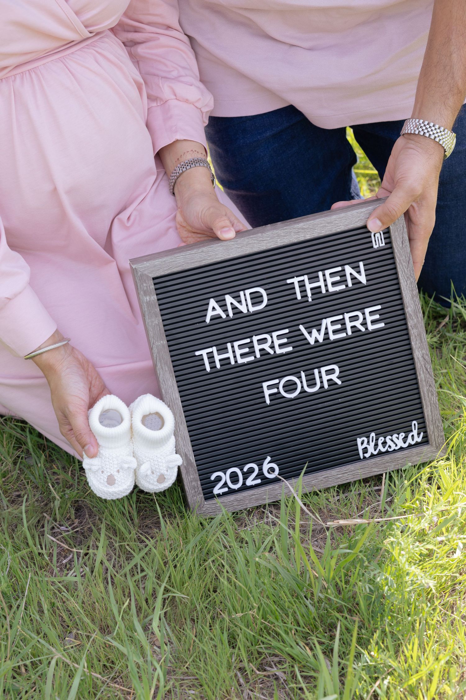 Couple holding baby shoes and a letter board sign reading 'And Then There Were Four 2026 Blessed' pregnancy announcement.