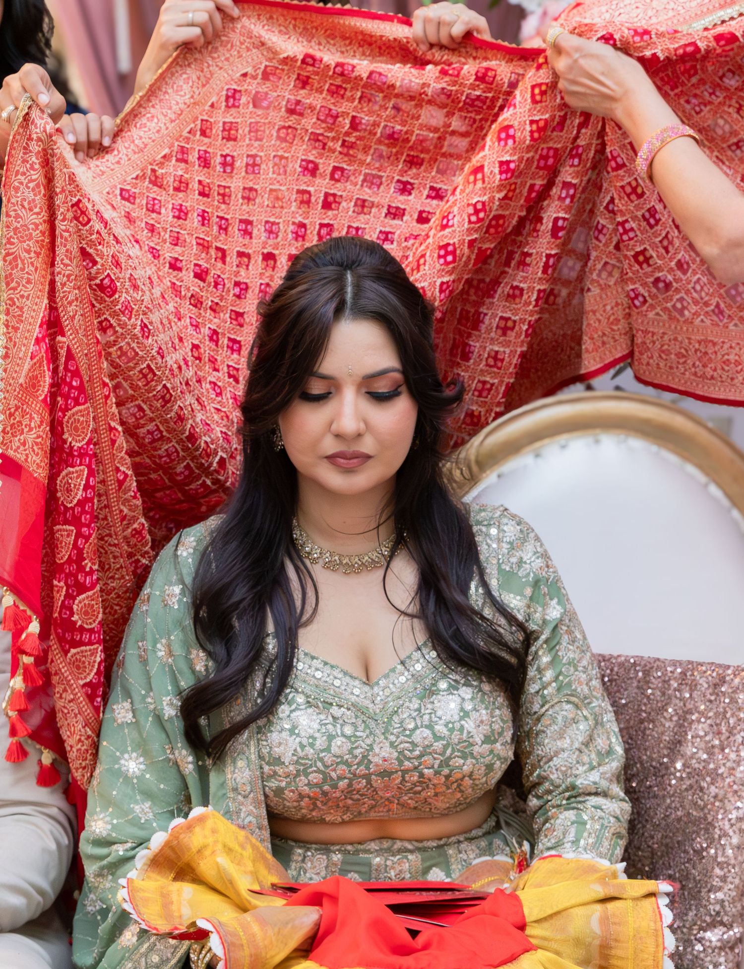 A woman in a mint green lehenga choli sits as others hold a red bridal dupatta behind her.