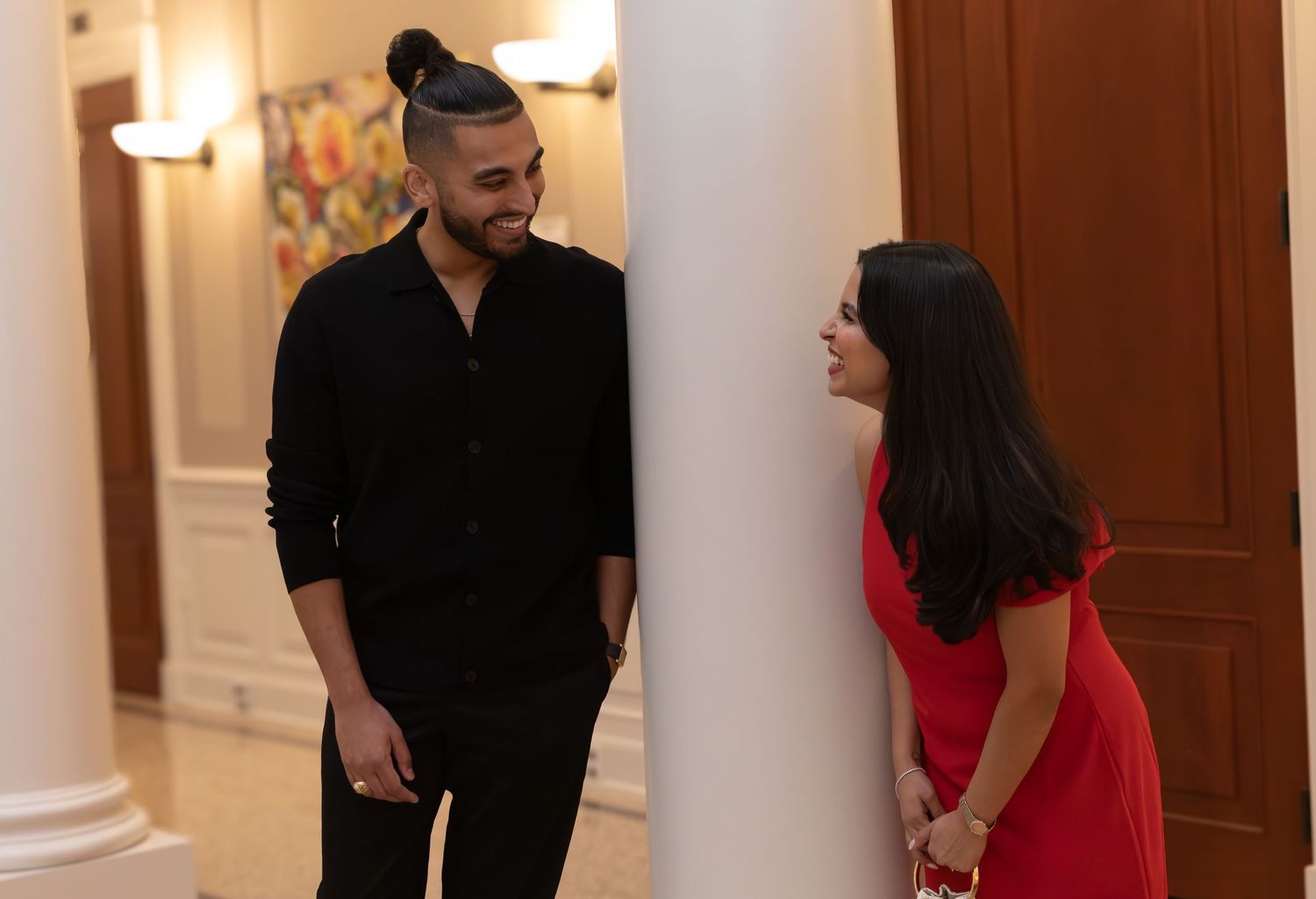 People in formal attire stand in an elegant hallway with white walls and warm lighting.