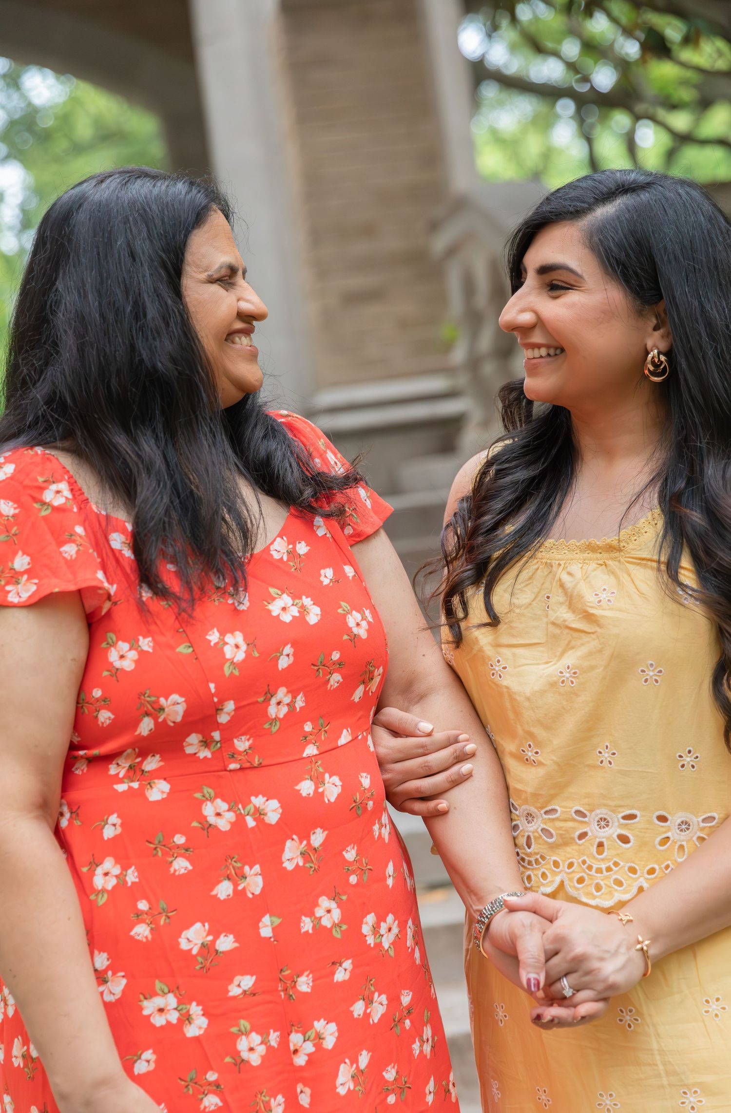 Two women wearing floral dresses in red and yellow share a joyful moment outdoors.