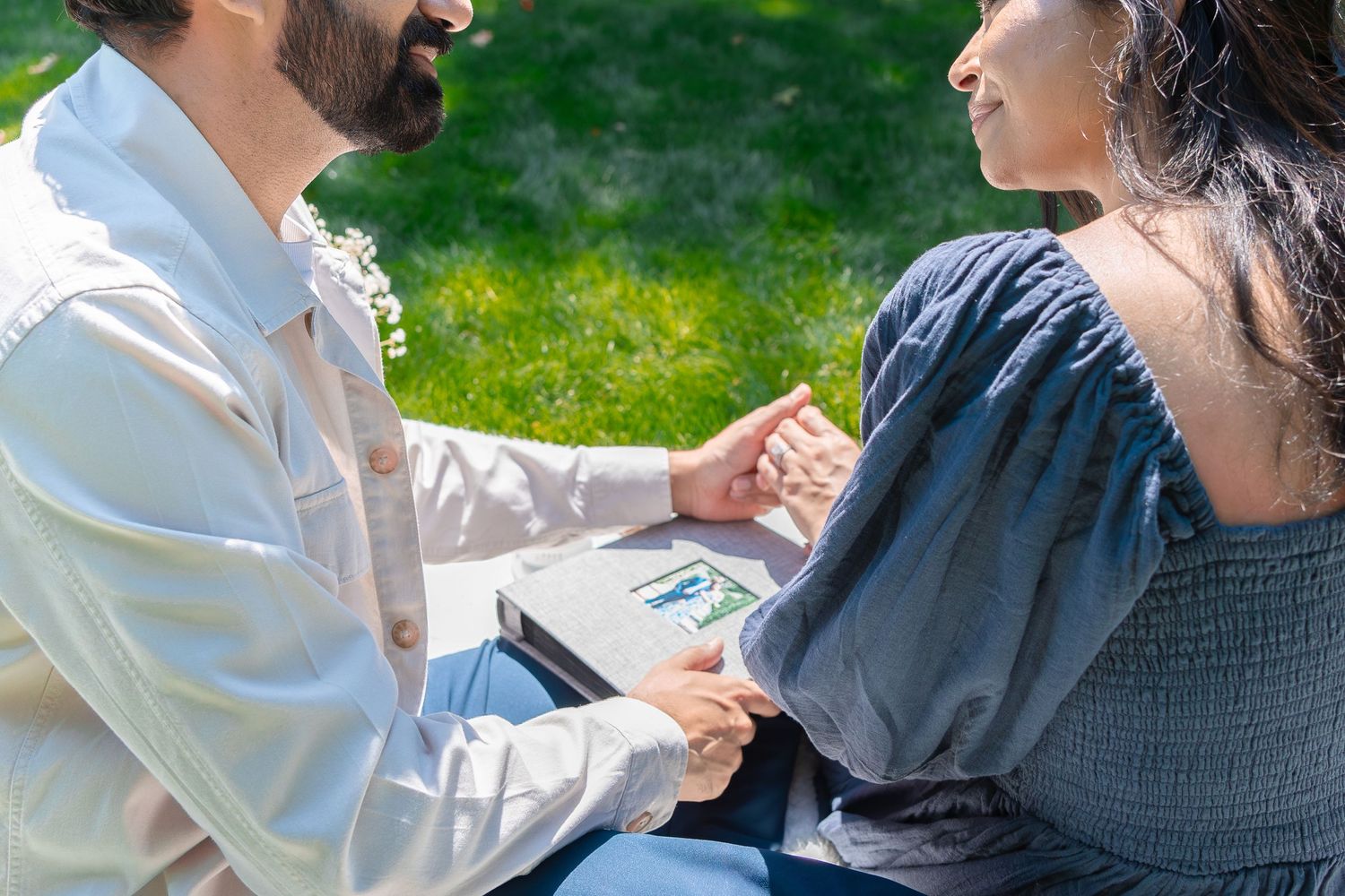Couple sitting outdoors holding hands on a sunny day with a photo album, sharing a romantic moment on green grass.